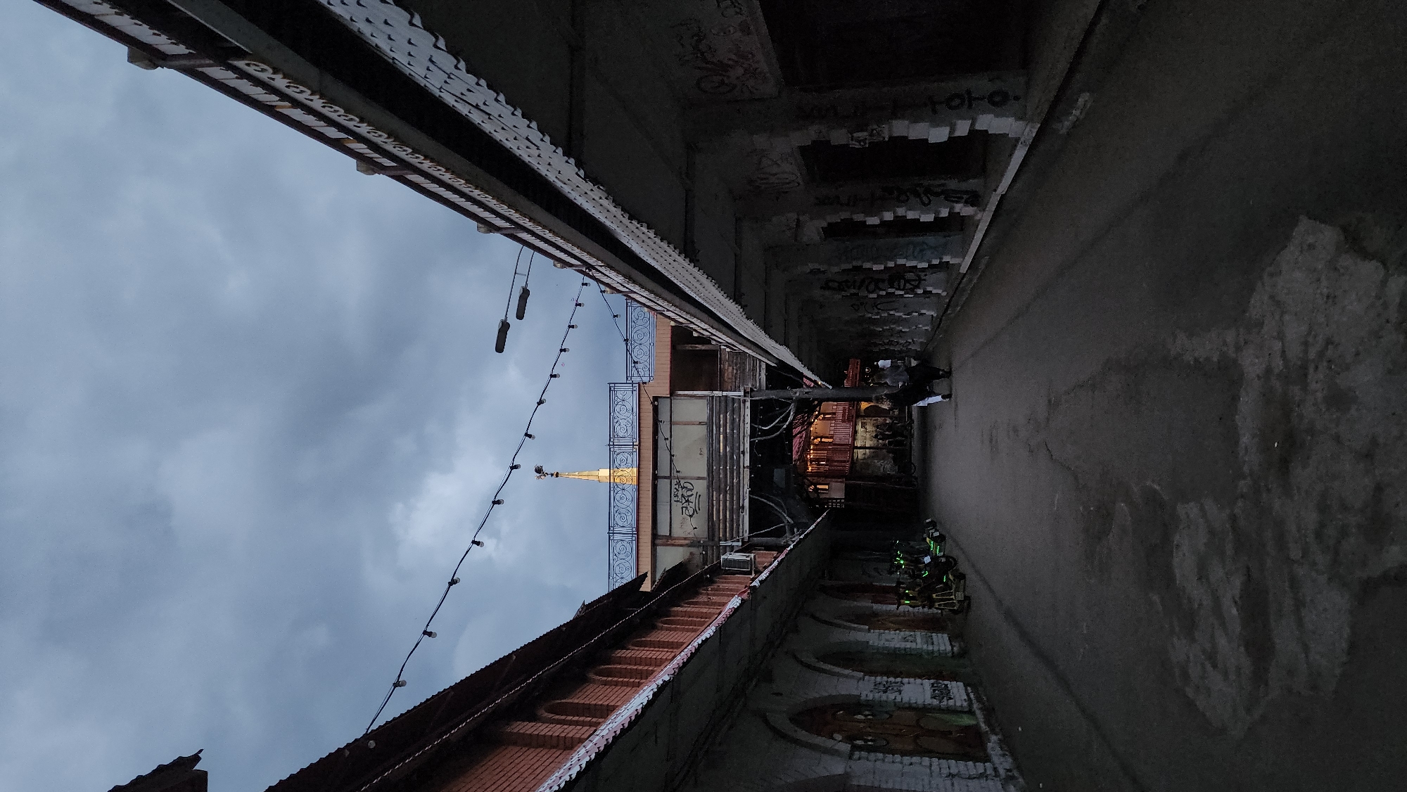 The photograph captures a narrow alley in Moscow, Russia, looking towards a gilded spire. The alley is bordered by buildings. The building on the left has a brick facade with arched windows. The building on the right is a gray concrete structure with graffiti on the upper walls. String lights crisscross above the alleyway. In the center background, a building with a gilded spire rises. Several people are standing in the alley near the background, under an overhanging structure. The sky above is overcast.