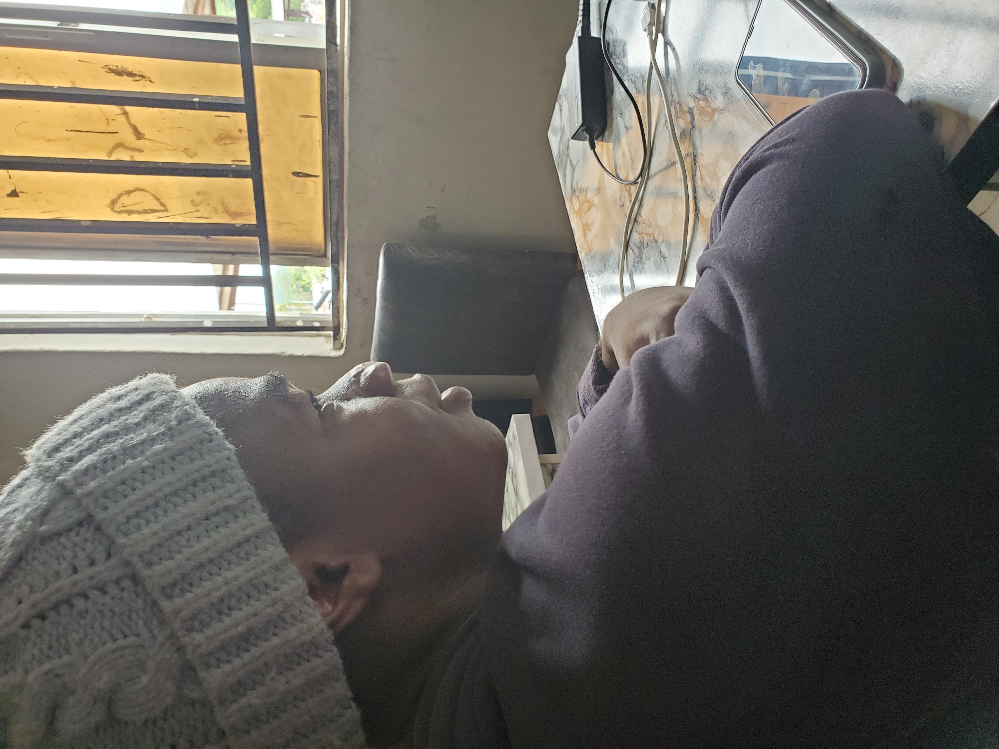 A person wearing a grey beanie and a dark purple hooded sweatshirt is lying on their back, looking towards a window with dirty yellow slats. The window is near a desk with a marble-like surface, where a black power adapter and white charging cables are located. Behind the desk, a dark grey chair is visible. The setting is likely an indoor space in Mararaba, Nigeria.