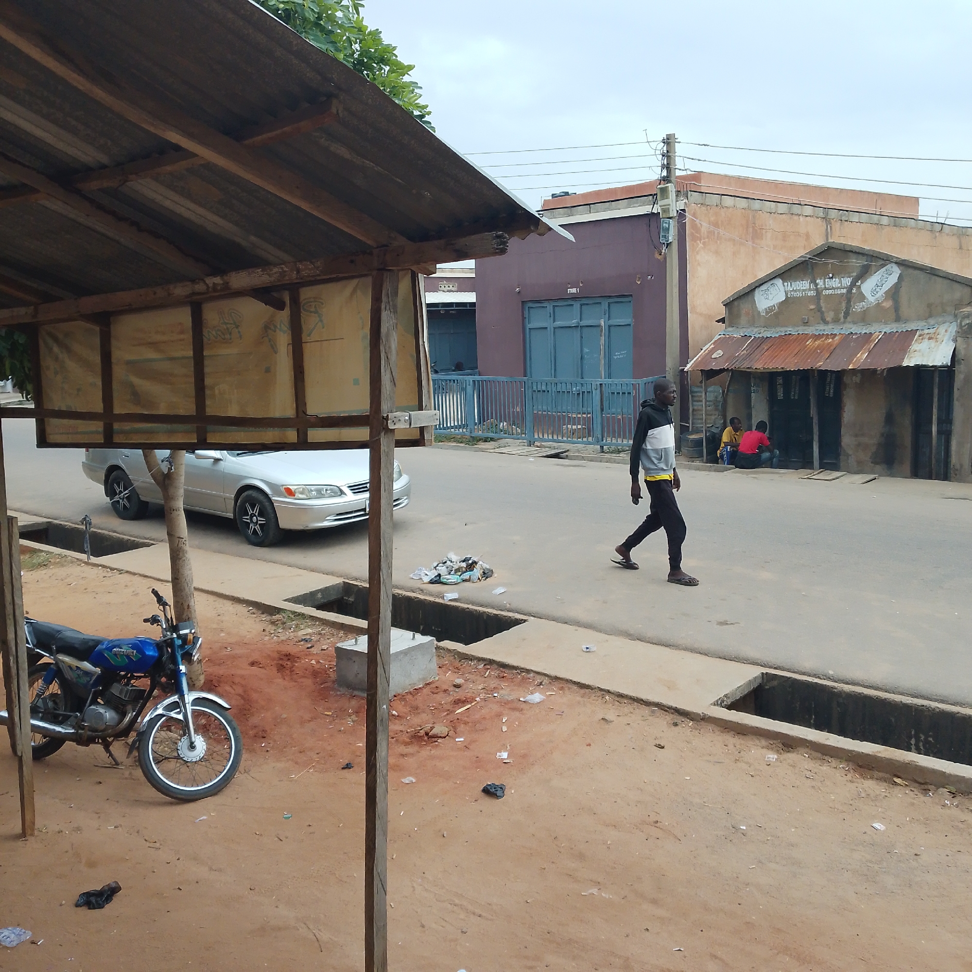 The image depicts a street scene in Pantame, Nigeria. In the foreground, a blue motorcycle is parked on sandy ground, with its front wheel turned. A wooden structure with a corrugated metal roof is partially visible on the left. A silver sedan is parked on the asphalt road. A person is walking across the street from right to left, wearing dark pants, a white and grey jacket, and a yellow undershirt. Further down the street, several people are seated in front of a building with a rusty corrugated metal roof. The buildings in the background have varying architectural styles and colors, with some featuring blue doors and windows. Overhead, power lines are visible against an overcast sky. The ground along the street includes a concrete gutter and dirt patches littered with debris.