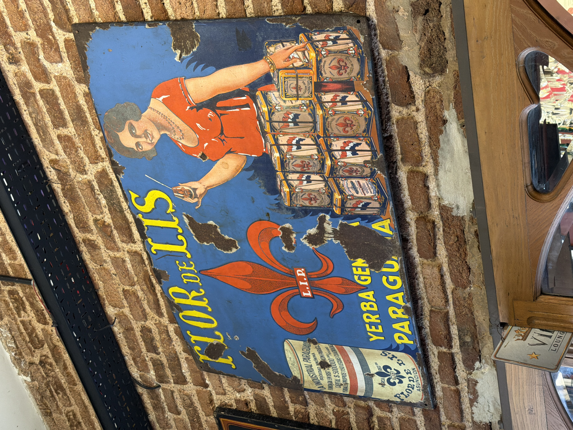 The image shows a worn and angled rectangular advertising sign for "Flor de Lis" Yerba Mate from Paraguay. The sign, mounted on a brick wall, features a woman in an orange dress stacking boxes of Yerba Mate. The backdrop is blue, and a large fleur-de-lis design is centrally positioned beneath the woman. The sign also includes text indicating the yerba mate is a product of "La Industrial Paraguaya Sociedad Anonima" founded in 1887. A small sign with "VII Lounge" is partially visible to the right of the image, indicating the potential location could be in a lounge or bar in Buenos Aires, Argentina.