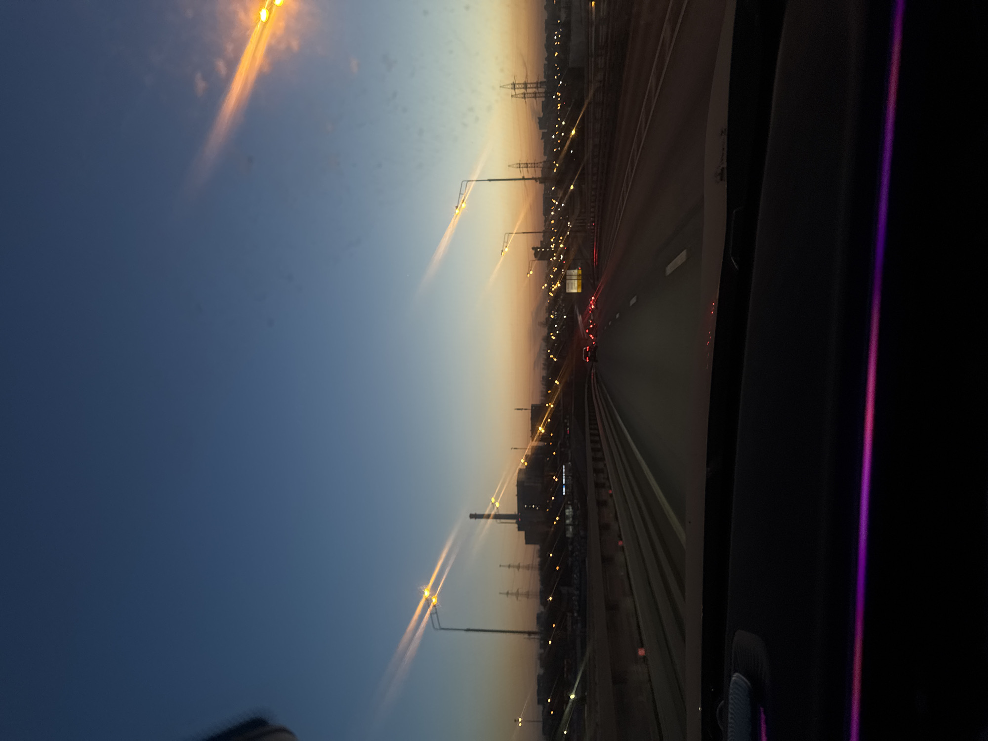 The image depicts a view from inside a vehicle traveling on a highway at dusk in Moscow, Russia. On the left side of the frame is a gradient sky, transitioning from a deep blue at the top to a lighter orange hue near the horizon. Streetlights line the highway, with their illuminated beams stretching upwards towards the sky. Visible along the right side are power lines, buildings, and infrastructure elements typical of an urban environment. The highway itself is multi-laned, and there are several vehicles visible with their taillights illuminated in the distance. In the foreground, parts of the vehicle interior, including the door trim, are visible.