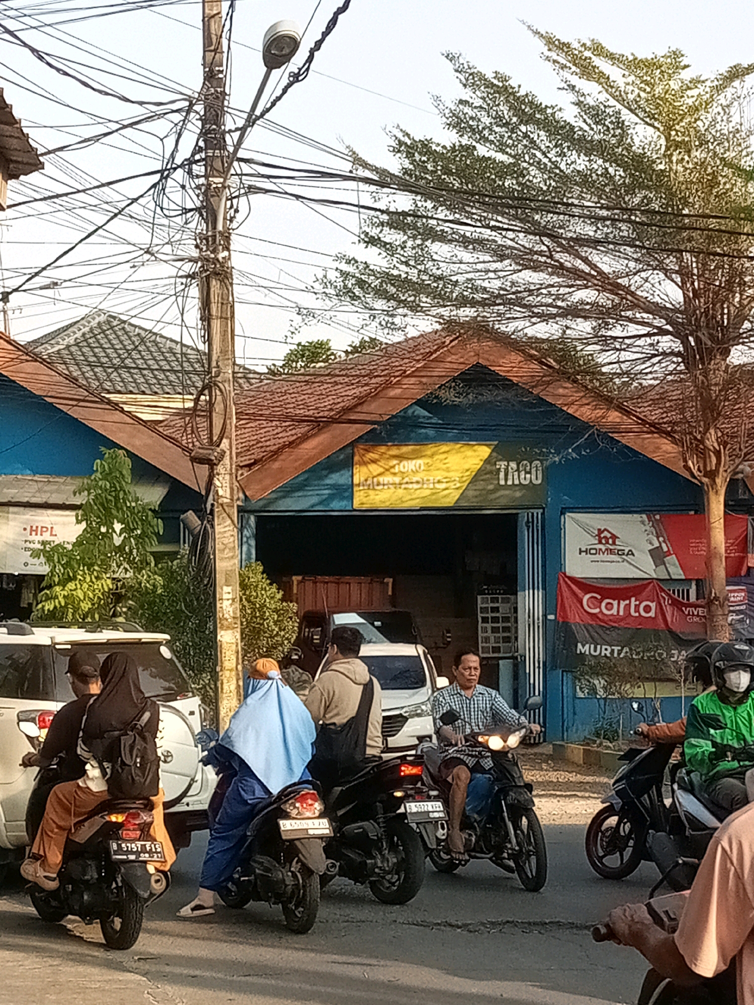 The image depicts a street scene in Tambun, Indonesia, likely during the daytime with a bright sky. Several motorcycles are present, with people riding on them. In the foreground, two motorcycles are stopped in traffic. The first motorcycle has a rider wearing a black hijab and an orange outfit, and a passenger behind them wearing a black hijab and backpack. The second motorcycle has a rider wearing a blue hijab and blue outfit. Further back, a man on a motorcycle wears a checkered shirt and jeans, and another rider in a green jacket and helmet is also visible.
Behind the motorcycles, there is a building with blue walls and a brown tiled roof. A sign above the entrance reads "TACO" in large white letters, with smaller text above and below. Several other signs are also visible on the building, including "HOMEGA" and "Carta". A large tree with sparse leaves is to the right of the building. A utility pole with numerous tangled wires is positioned in front of the building. In the background, more buildings with tiled roofs are visible, as well as a white car. The overall impression is of a busy street with traffic and commercial activity in a residential or commercial area of Tambun.