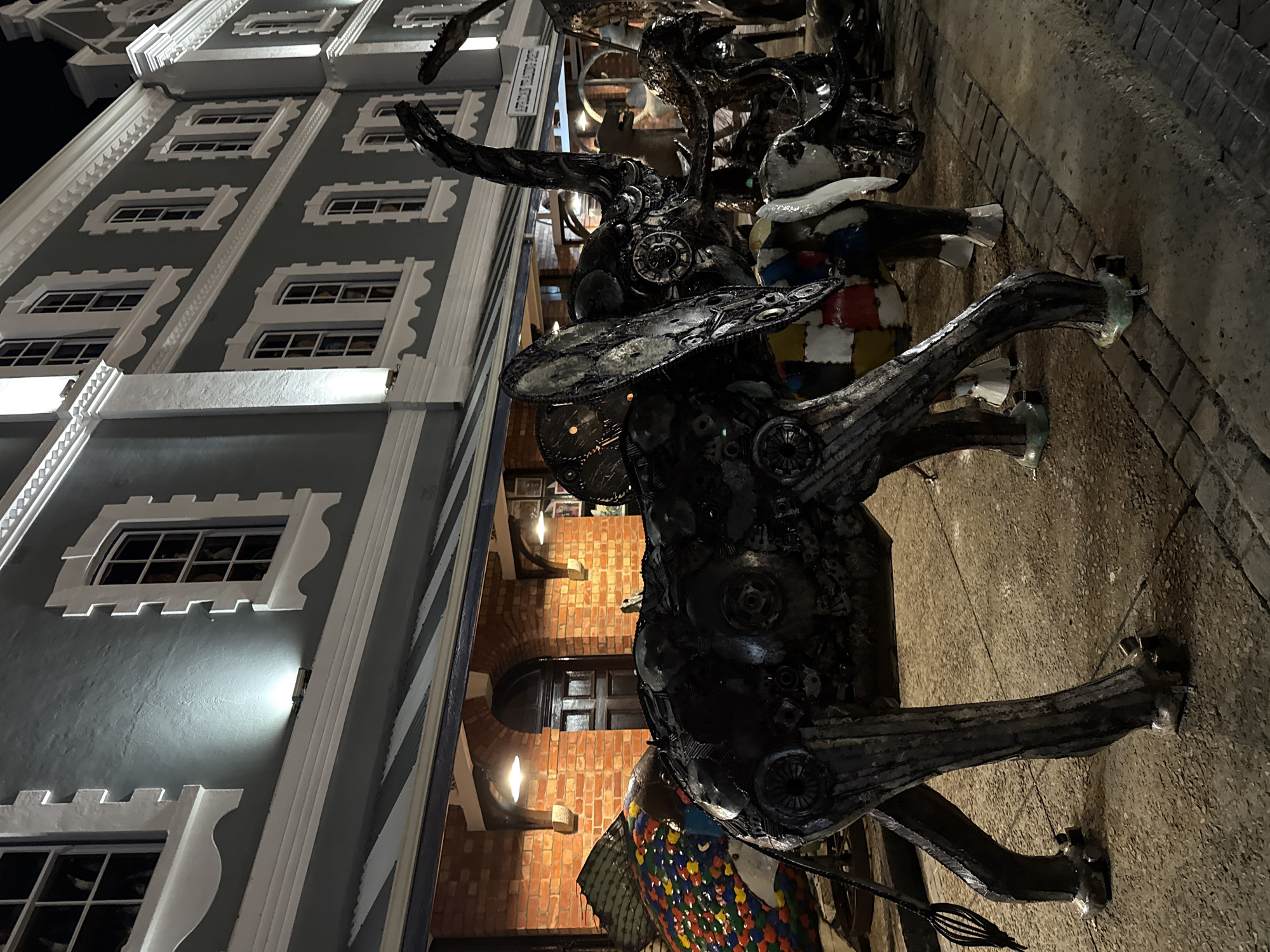 The image shows a large, metal sculpture resembling an animal, possibly a bull or a moose, standing on a sidewalk. The sculpture appears to be made from various metal parts and scrap materials. Behind the sculpture is a building with a distinctive facade, featuring gray walls and ornate white window frames and detailing. The background also shows a brick building and other architectural elements, including an arched doorway. The setting is identified as the city of Cape Town, South Africa. The lighting suggests it is nighttime. The sculpture is the primary focus, with the architectural backdrop providing context.