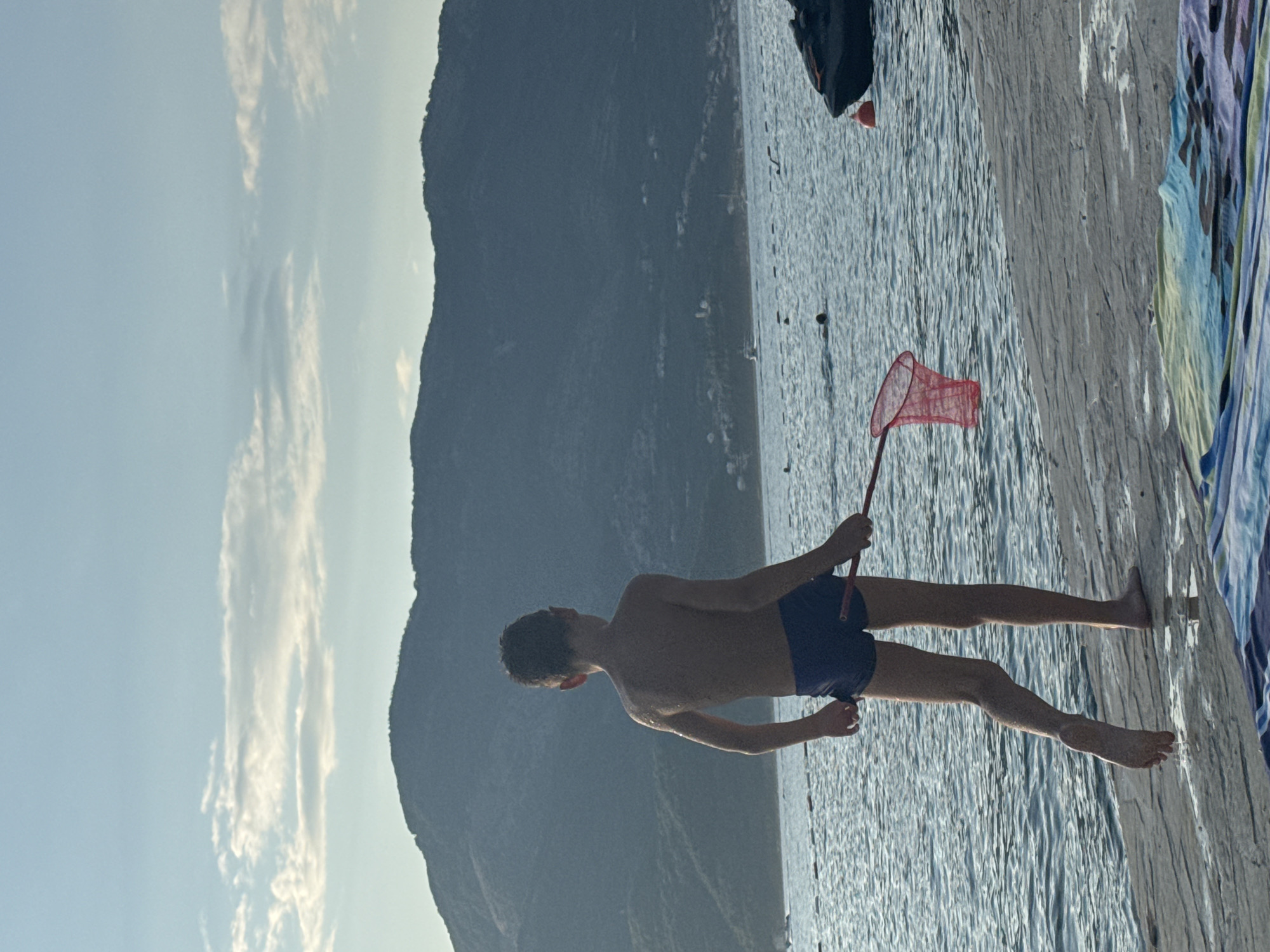A young boy stands on a paved surface near the water, facing away from the camera. He is wearing blue swim trunks and is holding a red net with a long handle. The boy's skin is wet, indicating he has likely been swimming. Behind him, the sea stretches out towards a large, mountainous landscape. The sky above is light blue with scattered clouds. In the distance, a jet ski is visible on the right side of the water, and other people are in the sea. A colorful towel is laid out on the paved ground in the foreground. The scene appears to be a coastal area.