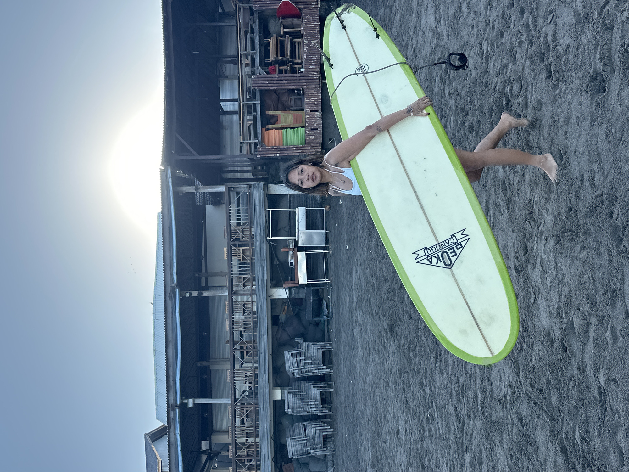 A young woman, positioned on the left side of the frame, carries a surfboard under her right arm. She is barefoot and wears a white bikini. The surfboard is light green with a white center and a black leash attached to its tail. Behind her, a building with a corrugated metal roof and wooden balconies stretches across the mid-ground. Stacks of chairs and tables are visible in front of the building. The ground is covered in dark sand. The sky is a pale blue, with the sun visible as a bright, diffused light source in the upper right quadrant of the image. The scene appears to be a beach in North Kuta, Indonesia, likely at sunrise or sunset given the quality of light.