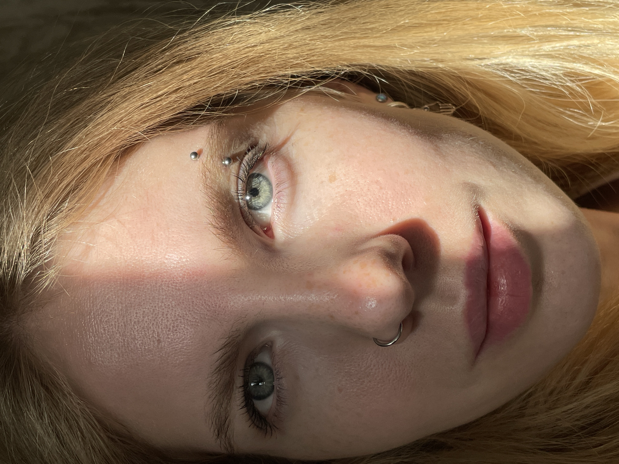This close-up, vertically oriented media file features a person's face, with a focus on their eyes, nose, and lips. The individual has fair skin with freckles visible on their nose. Their eyes are light-colored with long eyelashes. They have blonde hair, with some strands falling across their face.
Notable details include several piercings: one on the bridge of the nose, and two on the eyebrow. The lighting suggests it is daytime, with a strong light source casting shadows on the face, highlighting facial features and creating a dramatic effect. The scene appears to be indoors, possibly in a bedroom, given the soft texture of the background and hair. There is no visible text. The overall mood is contemplative and intimate.