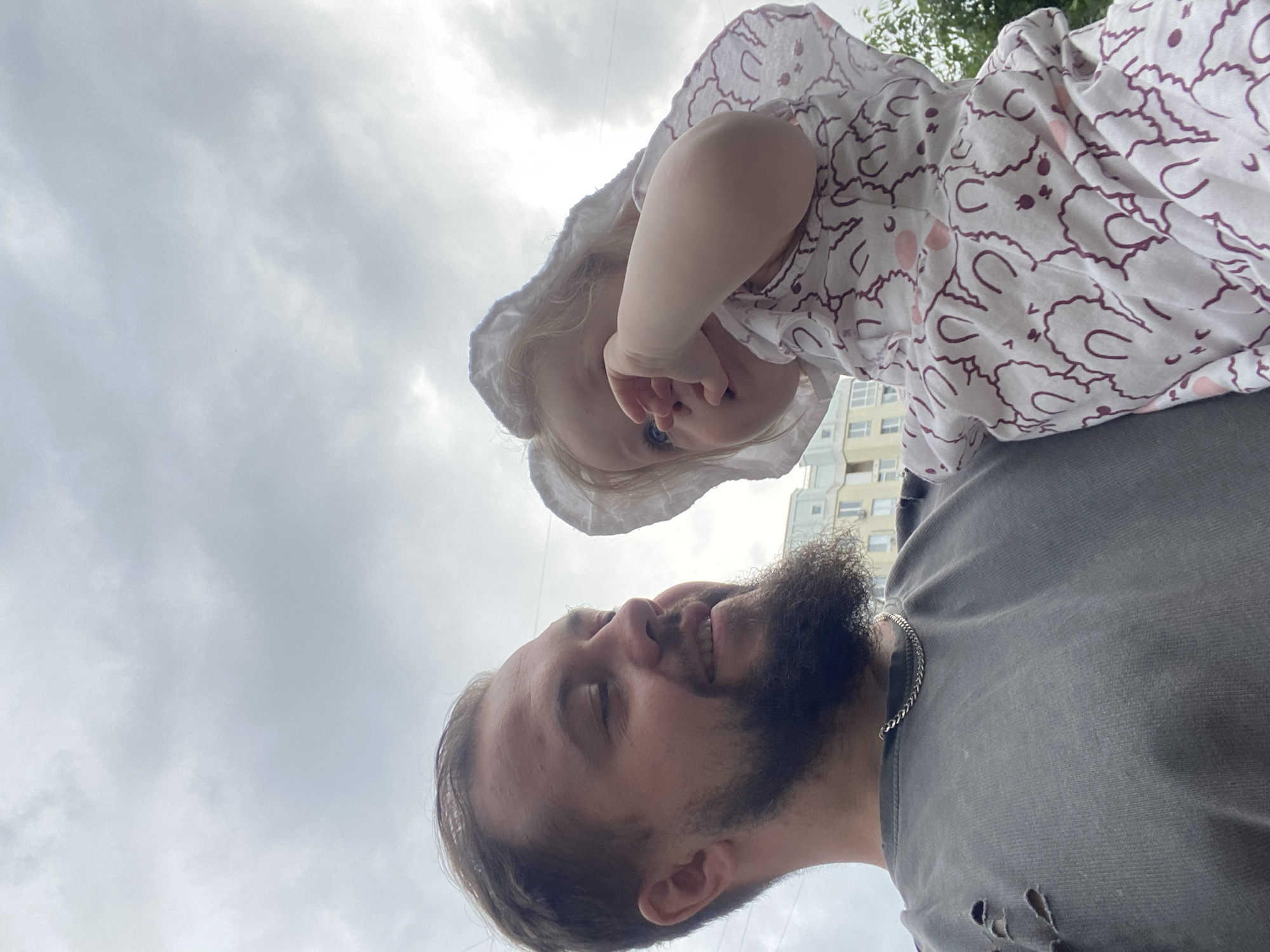 A low-angle outdoor medium shot shows a man holding a baby in his arms, with a cloudy sky as the background. The man, on the left, has a beard and is wearing a ripped gray t-shirt and a silver chain. He is looking to the right with a slight smile. The baby, on the right, is wearing a white sun hat and a white onesie with a pattern of cartoon bunnies. The baby's face is partially obscured by their hand, which is touching their face. In the background, a modern apartment building and green trees are visible. The overall setting appears to be urban, and given the context of the city of Vladimir, Russia, this scene likely takes place in a residential area or park.