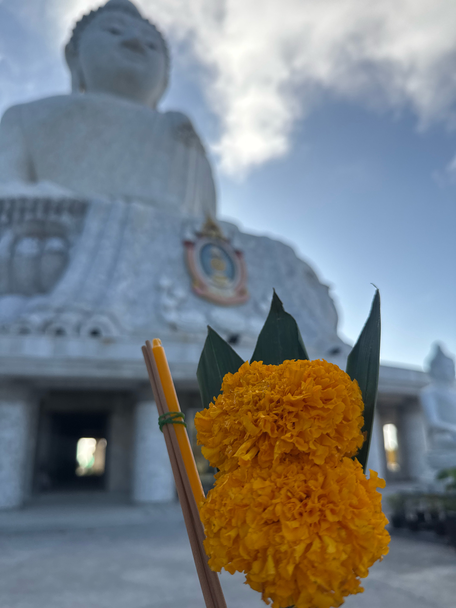 In Karon, Thailand, a close-up shot focuses on offerings in the foreground. Two bright orange marigolds are presented, nestled within dark green leaves and tied together with a thin green string. Adjacent to the flowers are two unlit incense sticks, one yellow and one brown. In the background, a large, white, blurred statue of Buddha is visible under a blue sky with scattered clouds. The statue appears to be situated on a paved area with architectural structures behind it. The scene suggests a place of worship or a cultural monument.