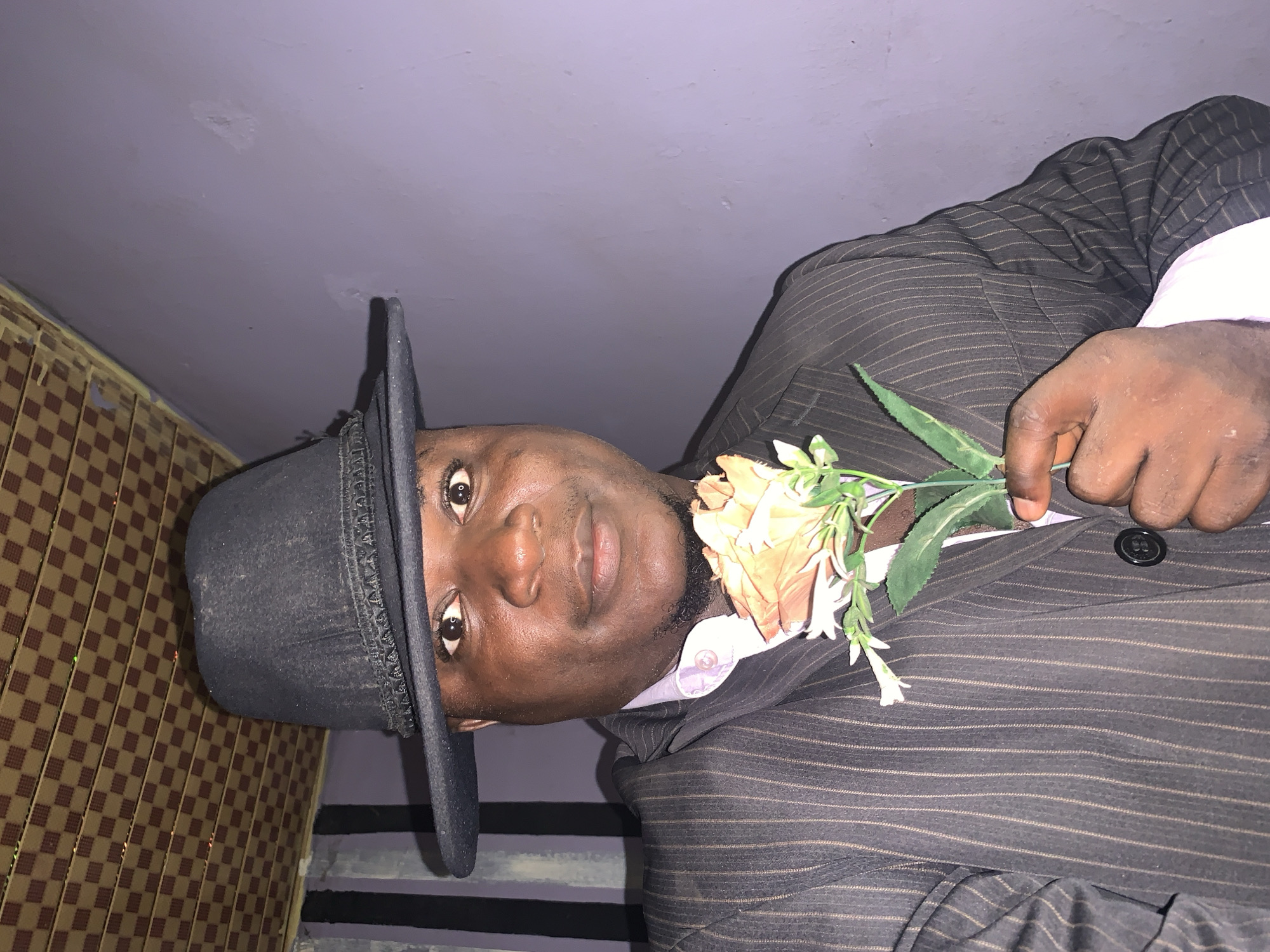 Akwanga, Nigeria: A dark-skinned man wearing a black hat and striped suit holds an artificial flower with orange and white blossoms. He is indoors, near a stairwell. A wall with a geometric pattern is visible to the left.