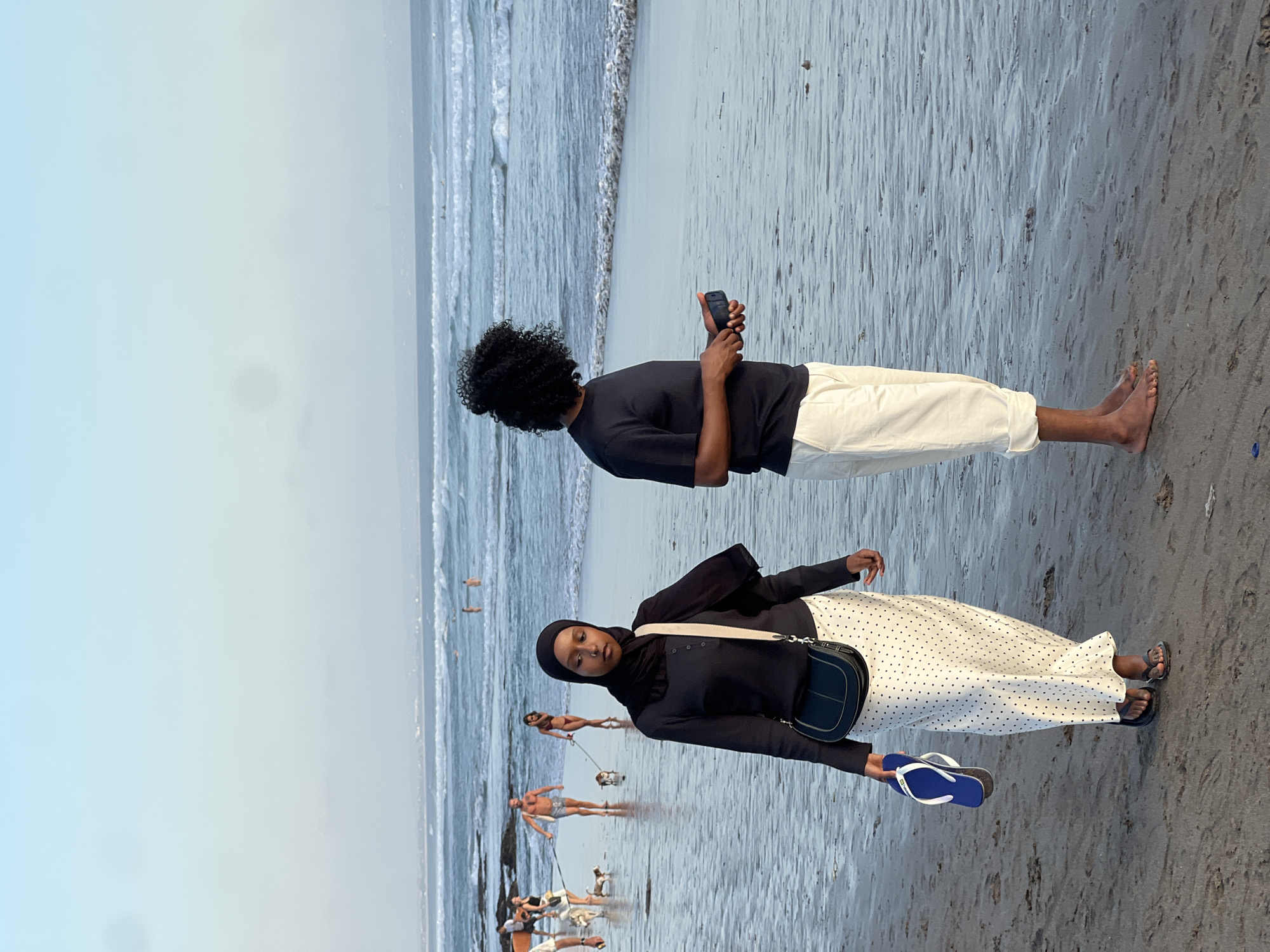 In North Kuta, Indonesia, on a beach with wet sand and gentle ocean waves, two individuals are depicted. In the foreground, a woman wearing a black hijab, a black long-sleeved top, and a white skirt with black polka dots walks towards the viewer, holding a pair of blue flip-flops in her left hand. She carries a black crossbody bag. To her right and slightly behind her, a person with curly dark hair, wearing a black t-shirt and off-white cropped pants, stands with their side to the viewer, looking out at the ocean and holding a black smartphone in their hands. In the midground, several people and two dogs are visible walking on the beach. The ocean extends to the horizon under a pale blue sky.