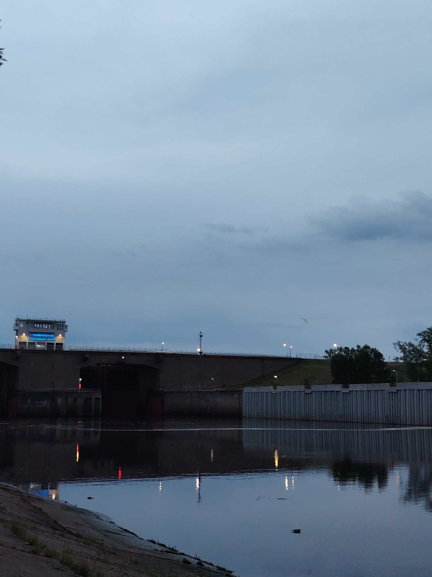 The image captures a low-angle view of the Chaikovsky Hydroelectric Lock, located in the city of Chaikovsky, Russia, under a cloudy, overcast sky. The lock structure, composed of dark grey concrete, spans across the frame. A control tower with illuminated blue signage rests on the dam. Below the main structure, the water reflects lights, including those on the dam and control tower. The shoreline is in the foreground. Vegetation can be seen on the riverbank.