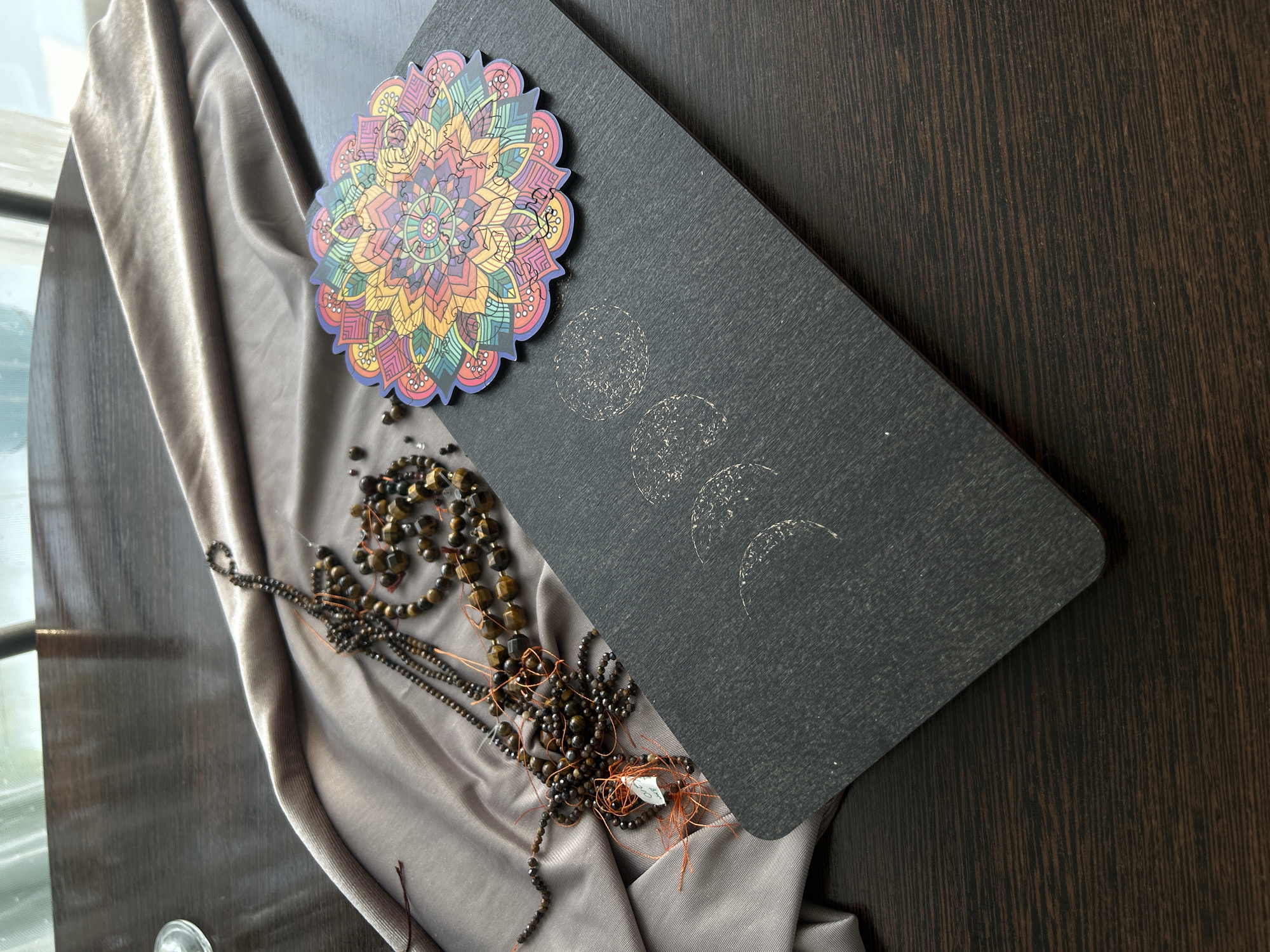 This is a close-up, overhead shot of a dark wooden table with various objects arranged on it. To the left, a piece of shimmery, taupe-colored fabric is draped. On top of the fabric, a tangled collection of dark, beaded necklaces lies. These beads appear to be a dark brown or black, with some larger, faceted tiger's eye beads interspersed. A small white price tag is attached to one of the necklaces with orange string. In the center right of the frame, a black rectangular mat is positioned diagonally. On this mat, a colorful, circular mandala sticker is placed. To the left of the mandala, on the black mat, there are faint, circular imprints that resemble phases of the moon. The background shows a window with sheer curtains and a glimpse of outdoor greenery. The overall setting suggests an indoor environment, possibly a home or a small shop. The image does not provide any specific details or context that would allow for identification of a location in Kemerovo, Russia.