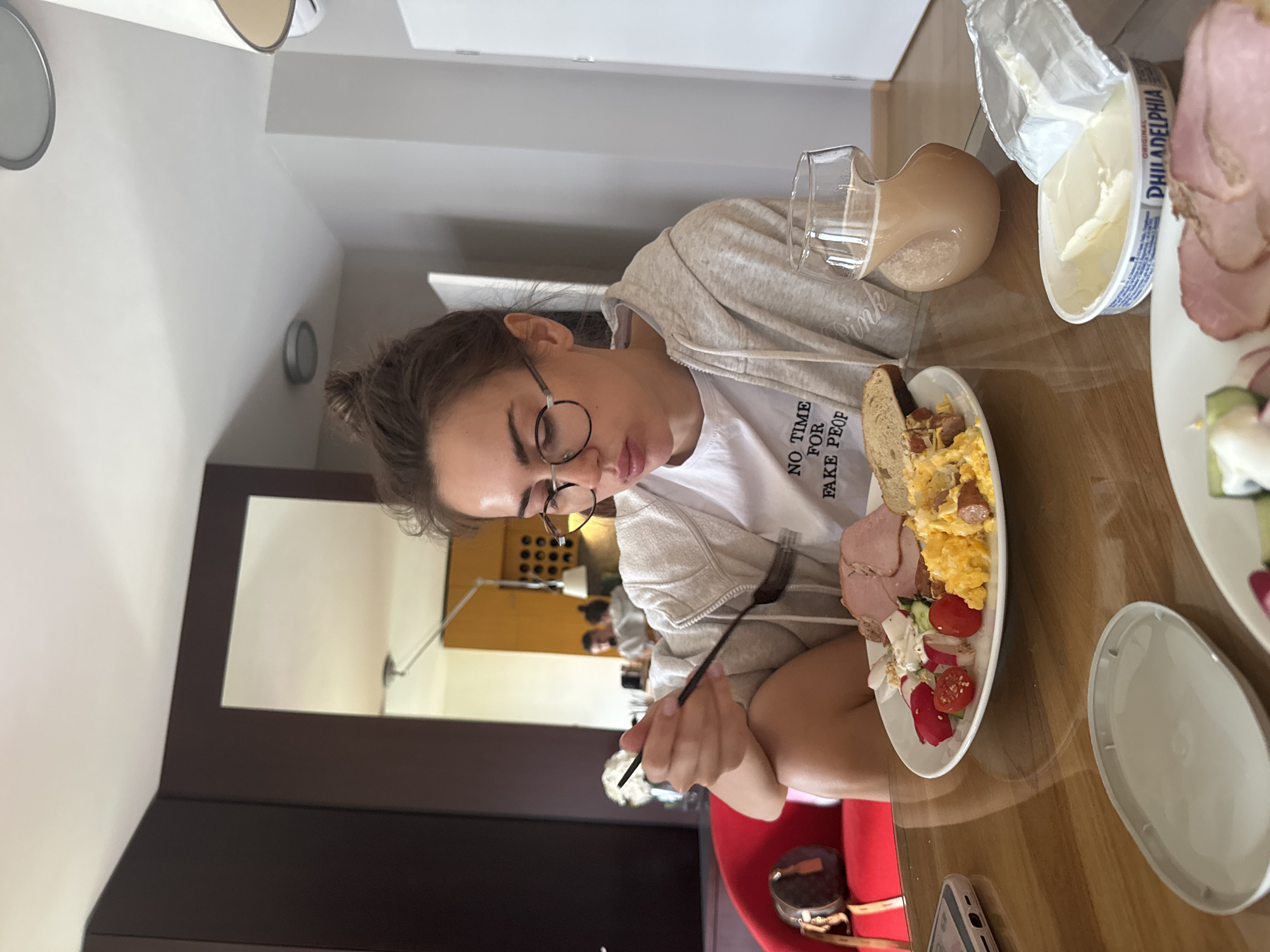An indoor scene, likely a dining area, shows a young woman seated at a wooden table, engaged in a meal. The image is rotated 90 degrees clockwise.
The woman, with brown hair tied in a bun and wearing glasses, is dressed in a white T-shirt and a grey hoodie. She is focused on her plate, holding a black fork. Her T-shirt has visible text that reads "NO TIME FOR FAKE PEOP", with the last word partially obscured.
On her white plate, there is a substantial breakfast or brunch spread: scrambled eggs with small pieces of sausage, a slice of bread, several slices of ham, and a small side salad containing cherry tomatoes and radish slices. Beside her plate, a clear glass jug holds a light brown beverage, possibly coffee with milk or a smoothie. An open tub of "PHILADELPHIA ORIGINAL" cream cheese is also visible on the table. To the right, another white plate holds more sliced ham. A smartphone in a light-colored case rests on the table near her hand.
The setting includes a wooden table and a red chair where the woman is sitting. In the background, a dark brown cabinet with a rectangular mirror reflects part of the room and what appears to be another person's silhouette in the distance. The lighting suggests daytime. The atmosphere is calm, with the woman concentrating on her meal.