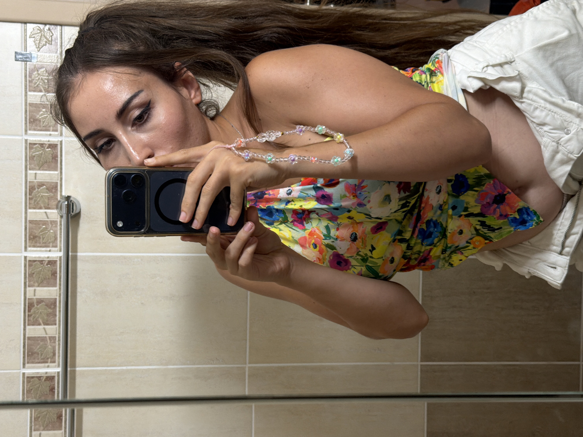 In Tersefanou, Cyprus, a woman with long, dark hair tied in a ponytail takes a selfie in a bathroom. She is wearing a colorful floral print top and white shorts. A beaded phone charm hangs from her right hand, which is also holding her phone. The bathroom features beige tiled walls and a silver towel rack.