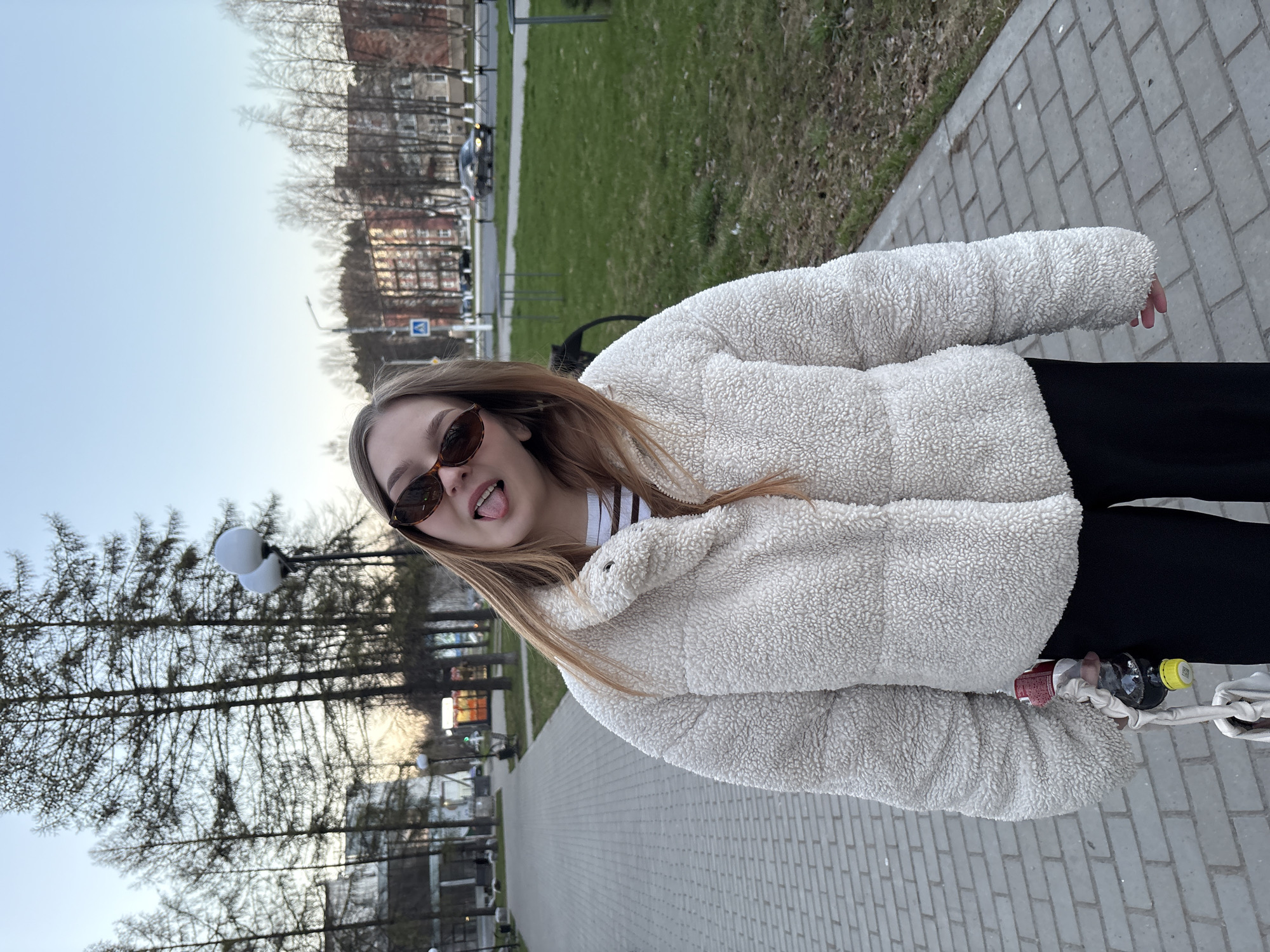 A young woman stands on a paved path with a brick pattern, likely in a park or urban area. She wears a white, puffy fleece jacket, black pants, and sunglasses. She sticks her tongue out, appearing playful.
In the background are trees, some without leaves, and a grassy area. Buildings are visible in the distance. Also in the background is a street with a car driving on it. Two spherical lamps are visible. A pair of plastic bottles are tied together and are also visible.
Given that the request specified Chaikovsky, Russia, it is assumed that this image was taken at a location in this city.