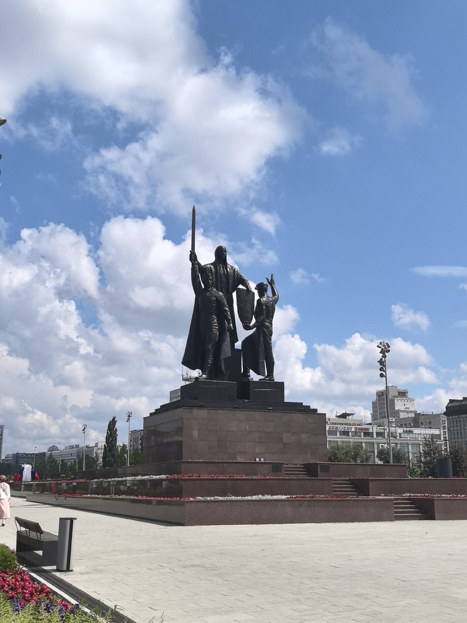 The image shows the monument "To the Fighters for Soviet Power" located in Perm, Russia. The bronze monument depicts three figures, with the central figure holding a sword aloft and a shield. Two other figures are positioned in front of and to the side of the central figure, one holding a sword and the other a shield. The monument stands on a multi-tiered granite pedestal. In the foreground, a paved plaza is visible, with a bench and a trash receptacle. Flower beds with red and pink flowers line the lower tiers of the pedestal. In the background, several multi-story buildings, trees, and streetlights are present, suggesting an urban environment. The sky is blue with scattered white clouds. A single person in a pink dress is visible walking on the plaza.