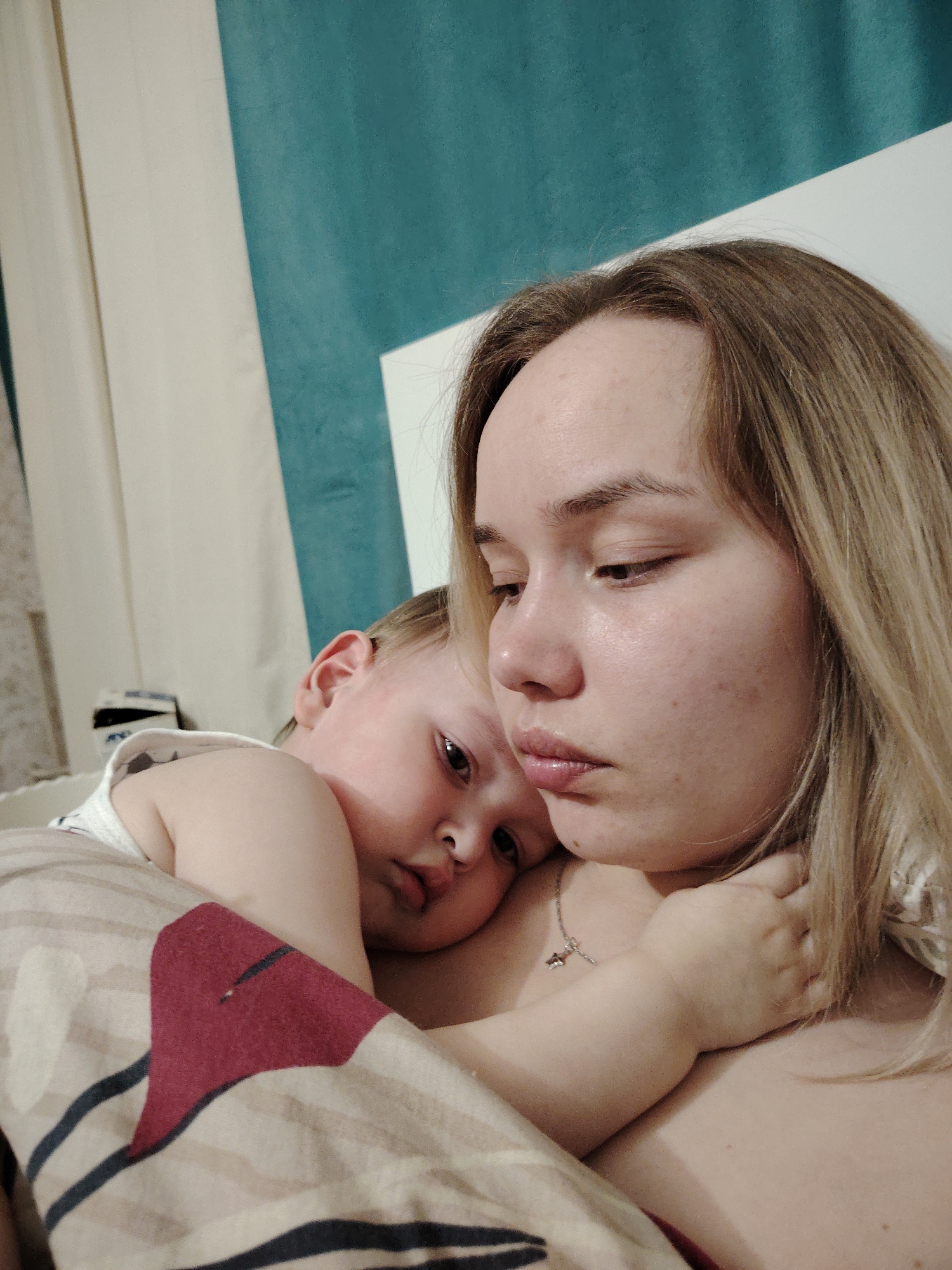 The image depicts a close-up of a woman and a child. The woman has blonde hair and is looking downwards with a somber expression. The child is nestled against her chest, appearing relaxed or sleepy. They are lying down under a striped blanket with a red heart shape. The background includes a wall with a two-toned blue and white color scheme.
There is no information in the image to determine the location is Марковский, Russia.
