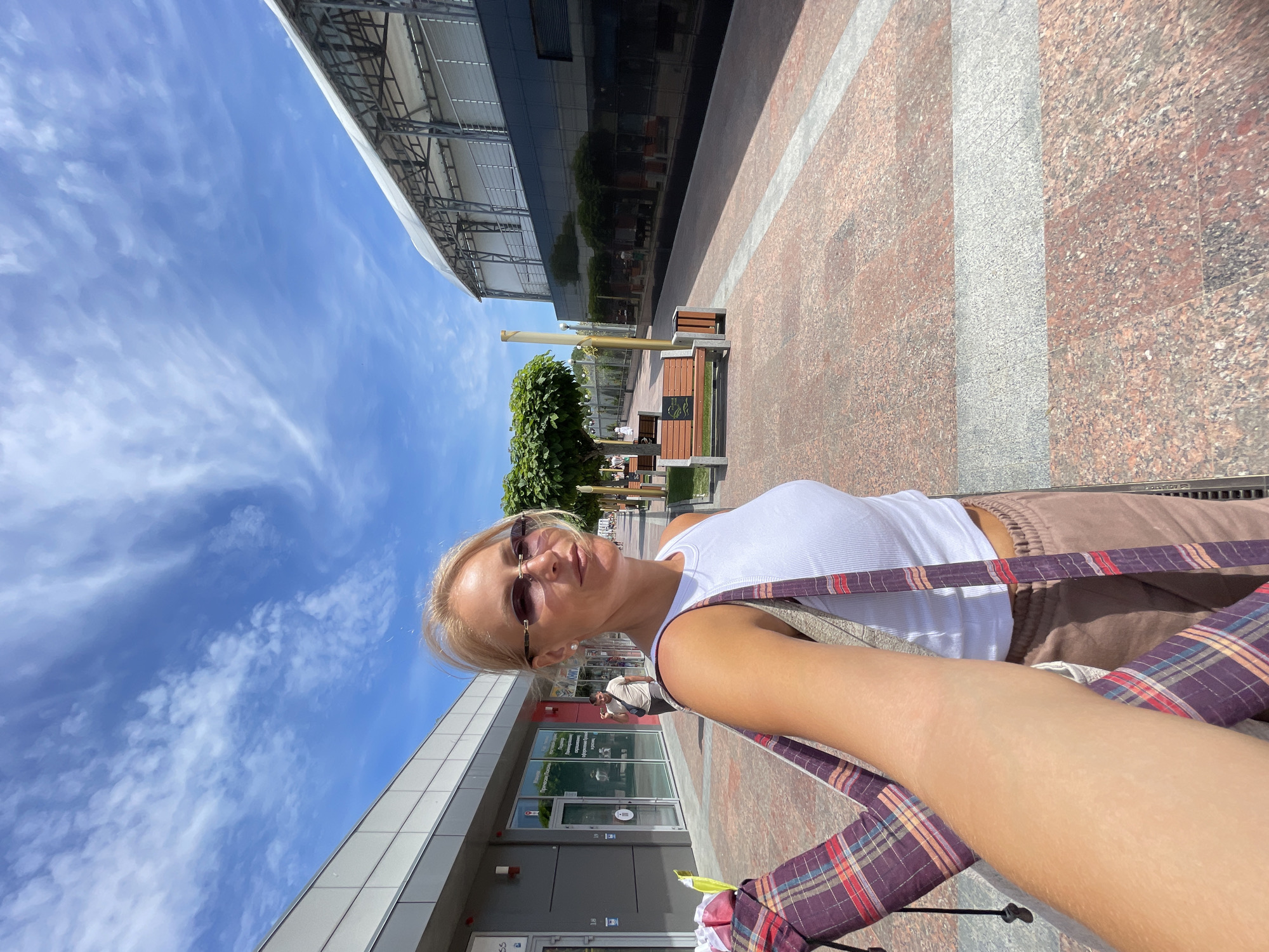 In this full shot, a woman is taking a selfie outdoors. She is wearing a white tank top and brown sweatpants, with a plaid bag slung over her shoulder. She is also wearing sunglasses. The background shows a modern building with large glass windows and a walkway lined with benches and trees. The sky is blue with scattered white clouds.