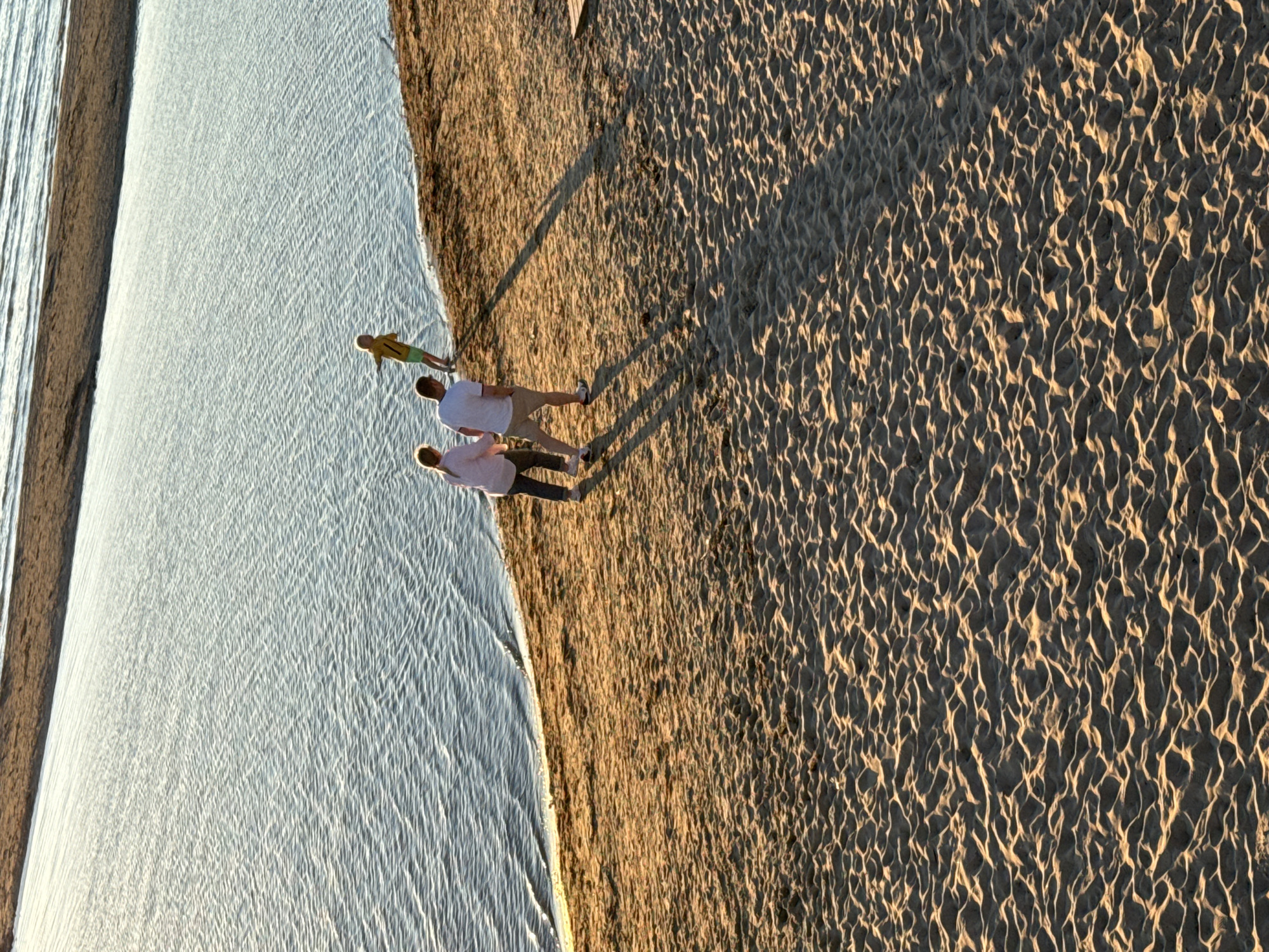 The image depicts a beach scene with three individuals near the water's edge. In the foreground, two adults are walking on the sand, with one wearing a light-colored shirt and the other a pink shirt and dark pants. To their right and further towards the water, a third person, appearing to be a child, is standing wearing a yellow shirt and green shorts. The background shows a large body of water with ripples, and a sandy shoreline. The lighting suggests it may be late afternoon or early evening. The environment is a sandy beach adjacent to calm water. It is not possible to determine the specific location within Tallinn, Estonia from the image alone.