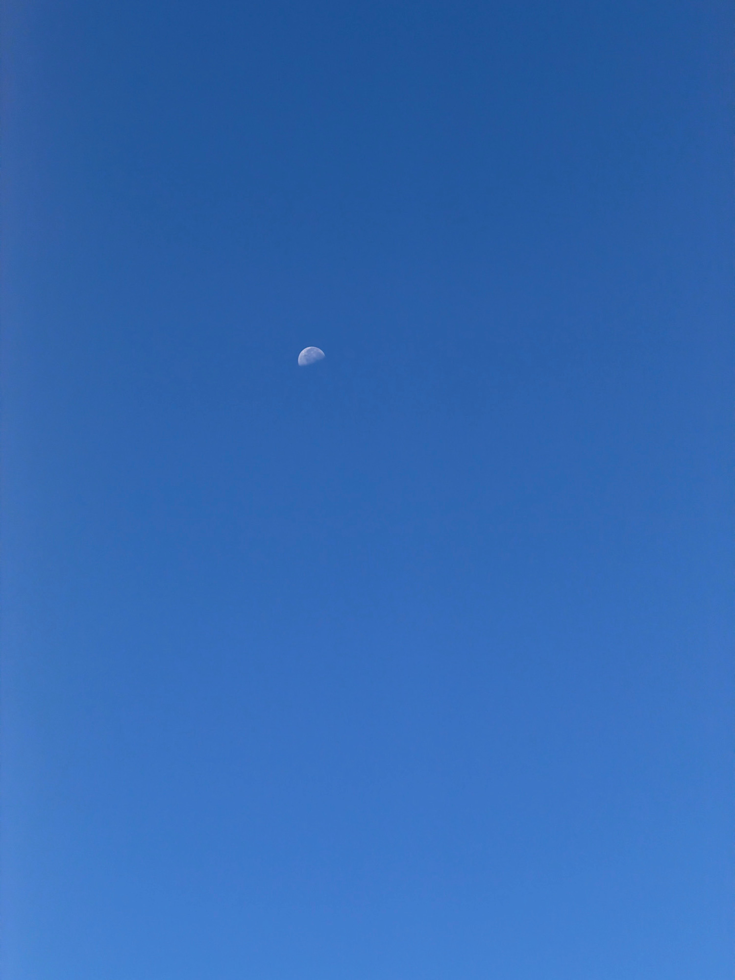 The image shows a clear, bright blue sky with a waxing gibbous moon visible in the upper center. There are no discernible landmarks or elements to indicate a specific location within the city of Arles, France, or any urban environment. The focus is solely on the celestial body against the vast expanse of the sky.