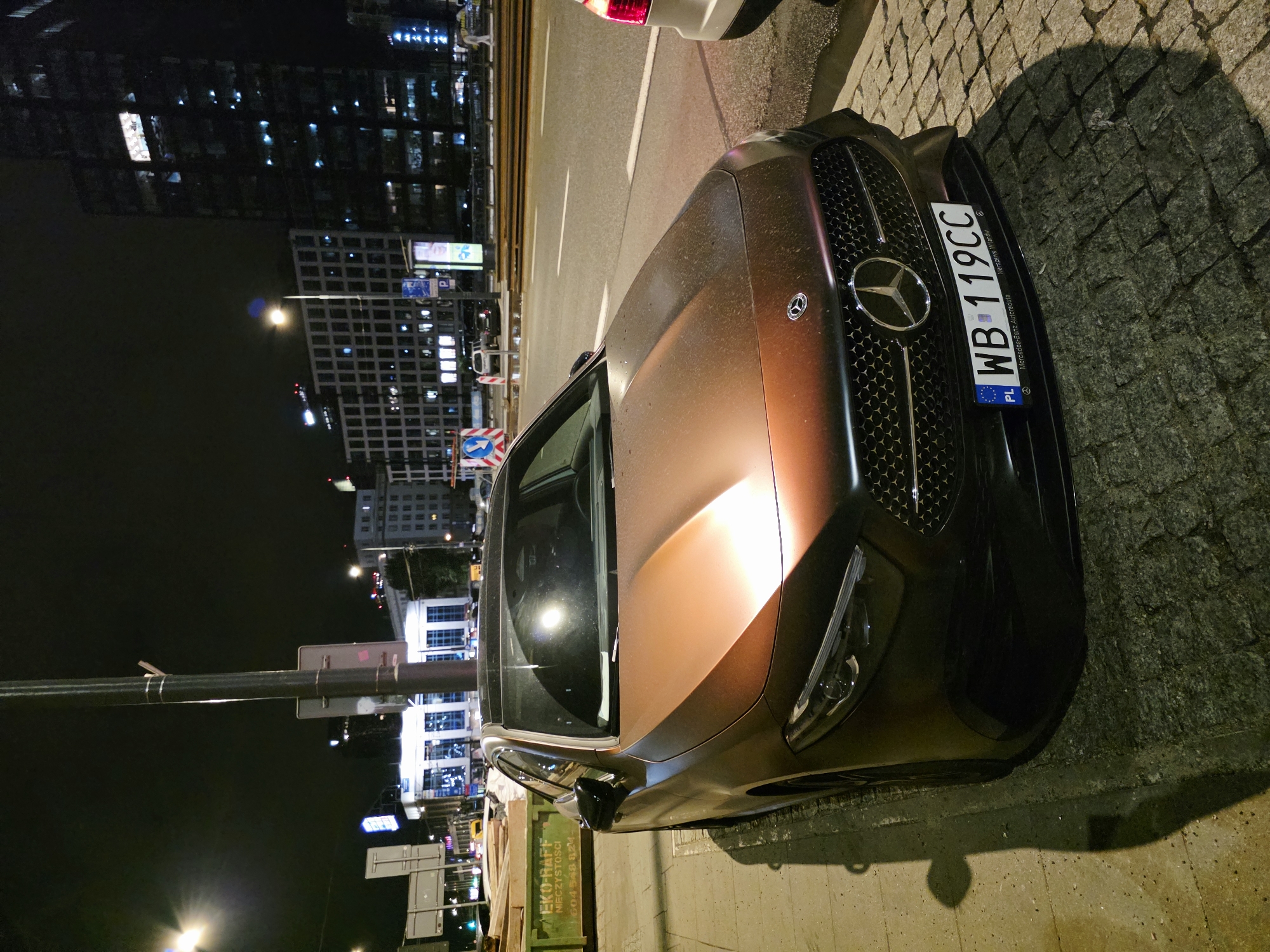 A low-angle, front-facing photograph captures a modern, bronze-colored Mercedes-Benz A-Class sedan parked on a cobblestone sidewalk at night in Warsaw, Poland. The car is illuminated by streetlights and the ambient glow of city lights. Its license plate, "WB 119CC," is clearly visible, with the "PL" designation indicating Poland. The car's grille features the prominent Mercedes-Benz star emblem. In the background, a multi-story building with numerous illuminated windows rises into the dark sky, hinting at an urban setting. Adjacent to the car, a bright green dumpster with white lettering, partially obscured, reads "EKO-RAFF NIECZYSTOŚCI" and a phone number starting with "604." The scene suggests a street-level view in a commercial or residential area of the city.