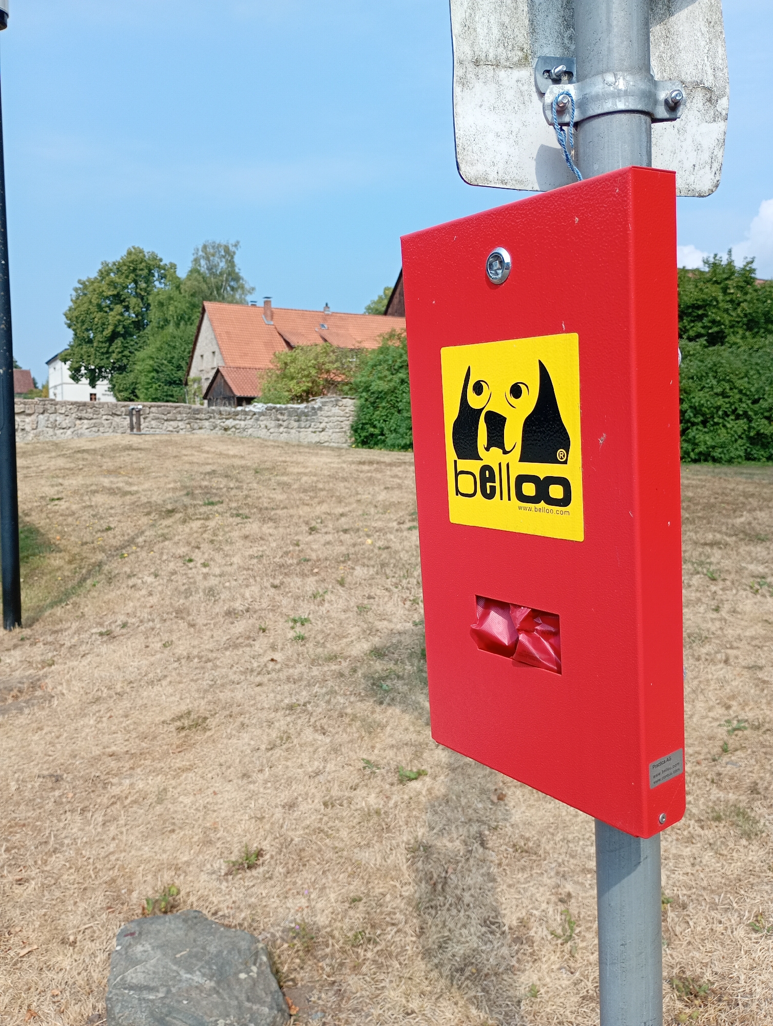 This is a close-up, eye-level shot of a red dog waste dispenser mounted on a metal pole in Walkenried, Germany. The dispenser features a yellow sticker with a black silhouette of two dogs and the word "belloo" in black lettering, along with a website address below. A slot at the bottom of the dispenser shows red plastic bags inside. The background consists of a dry, grassy field, a stone wall, and a few buildings with red-tiled roofs, suggesting a rural or semi-rural setting within the town. The sky is clear and blue. The image is taken outdoors during daylight.
