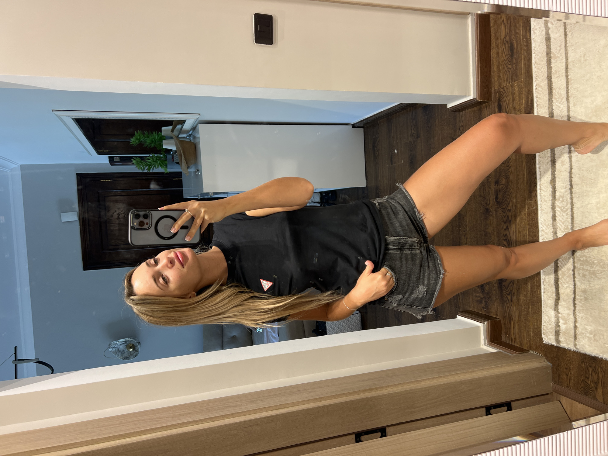 A young woman with blonde hair is taking a selfie in a room. She is wearing a black t-shirt with a red logo and distressed denim shorts. She is standing in front of a large mirror, which reflects her and the surrounding environment. The room appears to be a hallway or a dressing area, with wooden doors and walls. There is a dresser with plants and a hat in the reflection, and a rug on the floor. The lighting is soft, and the overall atmosphere is casual and relaxed. The scene is consistent with a residential setting in Sharm El Sheikh, Egypt, a popular tourist destination known for its beautiful beaches and resorts.