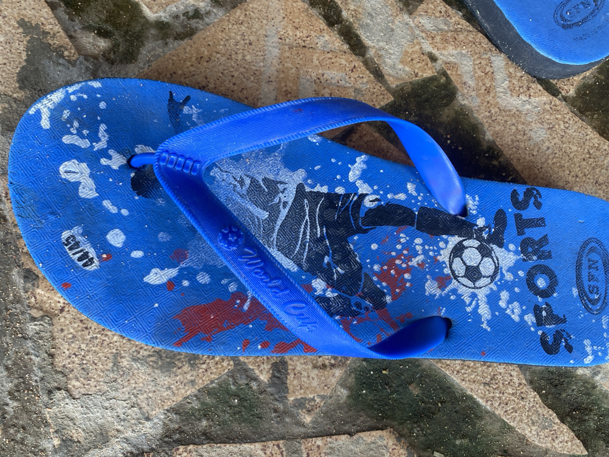 A blue flip-flop with a sports theme is lying on a textured surface that appears to be pavement or a tiled area. The flip-flop features a graphic of a soccer player kicking a ball, with the word "SPORTS" printed below it. There are also red and white splatters on the flip-flop, possibly indicating wear or a design element. A partial view of another blue flip-flop is visible to the right of the main subject. The environment suggests an outdoor setting. The location is identified as Jauro Yinu, Nigeria.
