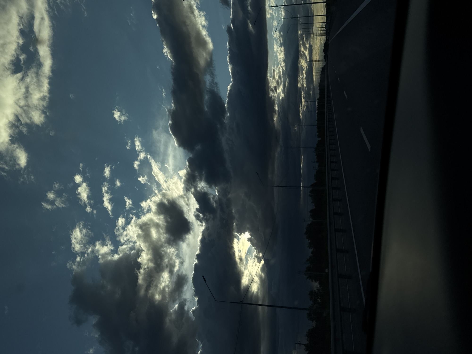 The image shows a view from a moving vehicle, possibly a car, on a highway or major road. The location is indicated as Sokolovyi, Russia. The dominant element is the sky, which is filled with a mix of dark, heavy storm clouds and patches of brighter, sunlit clouds. Streetlights line the side of the road, disappearing into the distance. A guardrail runs parallel to the road, separating it from a line of trees. The road itself is dark and appears to be wet. The inside of the vehicle is visible on the right edge of the frame, obscuring part of the view. There are no people visible in the image.