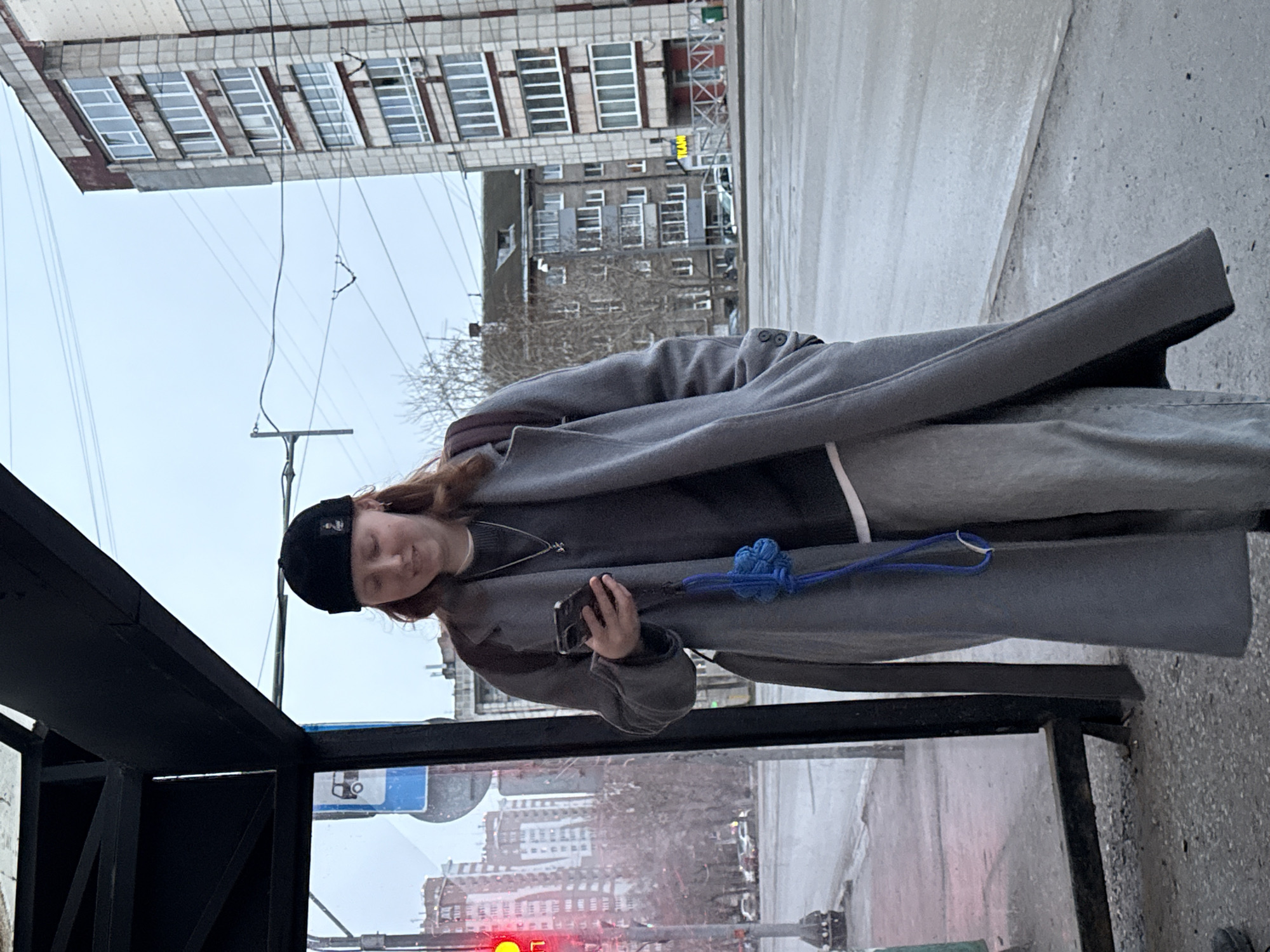 A young person in a black beanie, gray overcoat, and gray pants stands near a bus stop in Perm, Russia. They hold a cell phone in their left hand. A blue knotted object dangles by their side. The bus stop is constructed of black metal and glass. Outside the bus stop, a city street is visible with buildings, a traffic light showing red, and overhead wires. The sky is overcast.