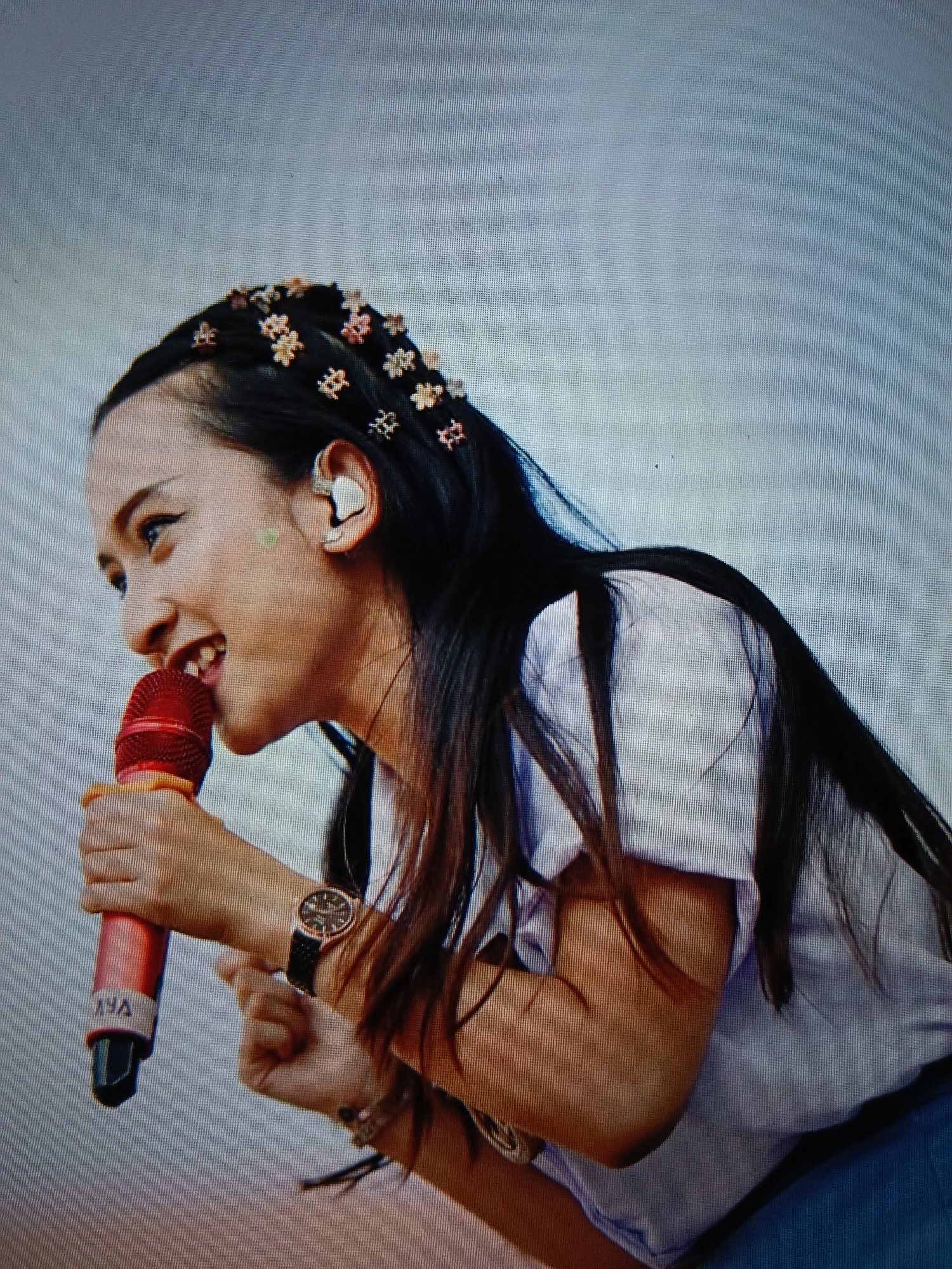 A young woman with long, dark hair, adorned with small, star-shaped hair clips, is seen in profile, holding a red microphone to her mouth. She wears a light-colored shirt and a watch on her left wrist. A light green heart-shaped mark is visible on her cheek. The background is a plain, light gray. The image appears to be a photograph taken indoors or outdoors with a neutral backdrop. There is no discernible information about the location in Raha, Indonesia, within the provided image.