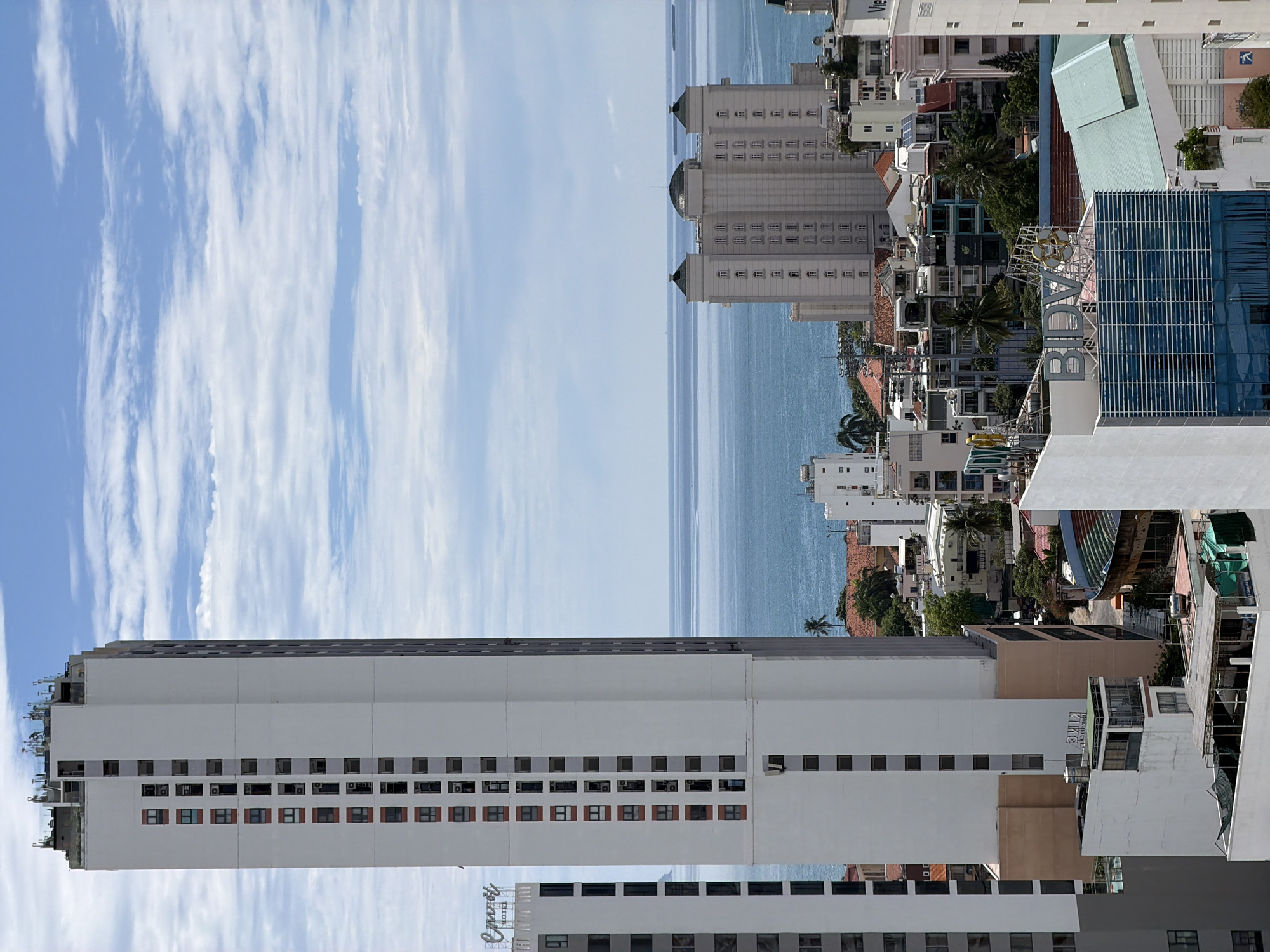 This vertically oriented image captures a bustling coastal cityscape, presumably Nha Trang, Vietnam, seen from a high vantage point, looking towards the sea. The bright blue sky, dotted with elongated white clouds, occupies the upper portion, suggesting a clear daytime view.
The scene is dominated by a dense array of urban architecture, featuring numerous high-rise and mid-rise buildings. In the prominent left foreground, a tall, slender white building stands out, characterized by rows of rectangular windows. White text on its lower section clearly spells out "Oceana Hotel." Further into the city, amidst a mix of residential and commercial structures, tropical vegetation, including palm trees, adds pockets of green.
Towards the mid-ground, a distinctive blue-tinted glass building with white vertical accents is visible, topped by a large yellow and white sign bearing the letters "BDV". Another building in this section displays the text "KMC". The architectural styles vary, from modern glass facades to more traditional, multi-storied concrete structures with tiled roofs.
Beyond the urban sprawl, the vast, shimmering blue expanse of the ocean stretches towards the horizon, merging with the lighter blue of the sky in the distance. Along the coastline, more buildings are clustered, including a complex of light-colored structures with domed or pointed roofs, possibly part of a resort or hotel. The overall impression is one of a vibrant, developed coastal city. The weather appears fair and sunny, indicating a pleasant day. No people are discernible in the scene.