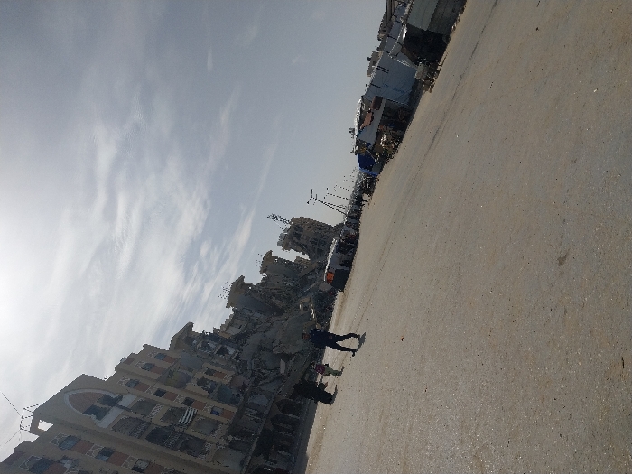 The image displays a wide, paved street or square in Khan Yunis, Palestinian Territory, under a bright, partly cloudy sky. On the left side of the frame, multiple multi-story buildings are visible, severely damaged with extensive structural collapse, exposed rebar, and visible debris. One prominent building on the left exhibits distinct architectural arches on its upper floors, now compromised. In the left midground, three individuals are present on the paved surface: one figure in dark clothing, a smaller figure in light attire, and a third individual in dark blue clothing, all walking. Along the right side of the street, a series of makeshift structures constructed from corrugated metal, tarps, and other materials line the thoroughfare, appearing as temporary market stalls or shelters. Streetlights extend into the distance along the right side of the road.