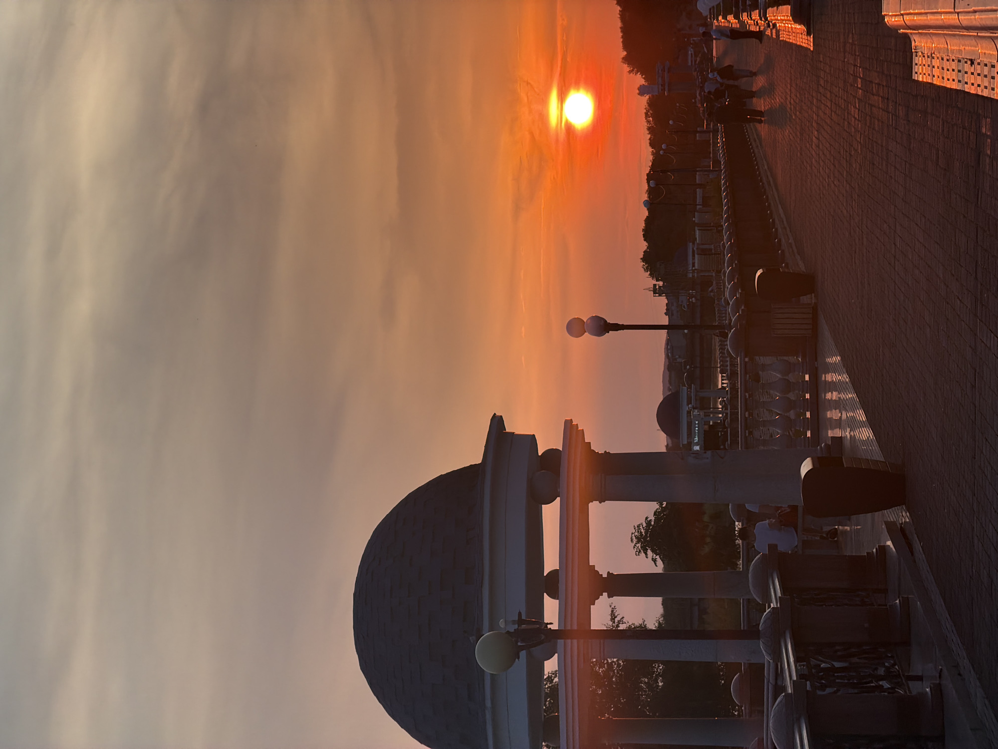 The image depicts a sunset scene on a paved walkway lined with lampposts and ornate railings. In the foreground, a domed structure with columns is visible. Several people are scattered along the walkway, some walking and others sitting. The sun is low on the horizon, casting a warm orange glow across the sky and illuminating the scene. The light reflects off the tiled walkway, creating a golden path. Trees are visible in the distance, silhouetted against the bright sky. The overall atmosphere is peaceful and serene, likely captured during the late afternoon or early evening. The location is identified as Birobidzhan, Russia.