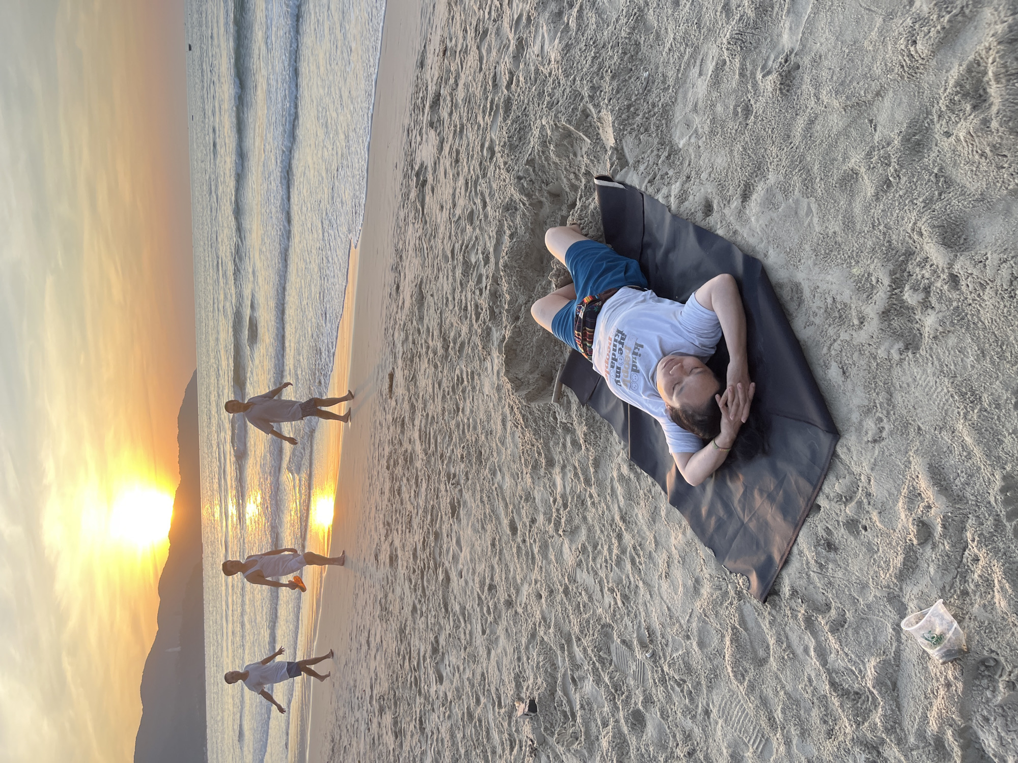 The image depicts a serene beach scene in Đà Nẵng, Vietnam, during sunset. In the foreground, a person lies on a black mat on the sandy beach, resting with their eyes closed and hands behind their head. They are wearing a white t-shirt with text and blue shorts. Further back, three men are walking along the shoreline. The sun is setting over the ocean, casting a warm golden glow on the water and the sky, with mountains visible in the distance. The waves gently lap at the shore, and the beach is speckled with footprints. A discarded plastic cup lies on the sand near the person on the mat.