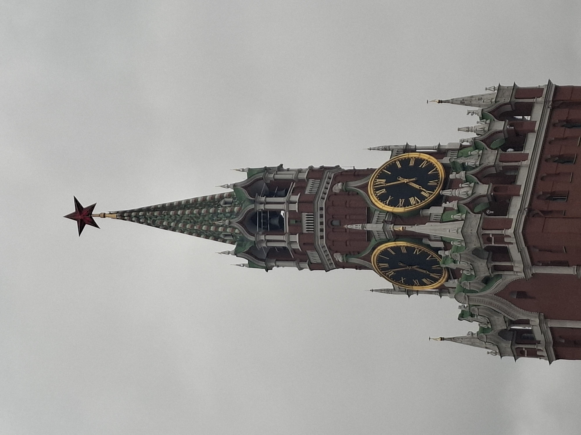 The image captures a close-up view of the upper section of the Spasskaya Tower, part of the Moscow Kremlin in Moscow, Russia, against a plain, overcast sky. The image is rotated 90 degrees clockwise from its natural upright orientation.
The prominent architectural landmark features two large, circular clock faces, both adorned with golden frames and black dials. Golden Roman numerals (I, II, III, IV, V, VI, VII, VIII, IX, X, XI, XII) mark the hours, and the hands on both clocks point to approximately 10:45 or 10:50.
Above the clocks, the tower transitions into an ornate spire. The spire has a distinct pattern of green and darker shades, tapering upwards to a pinnacle. At the very top, a prominent, five-pointed red star is affixed, a recognizable symbol of the Kremlin towers. The main structure of the tower visible in the shot consists of reddish-brown brickwork, accented with intricate grey and white decorative elements, balustrades, and smaller spires or finials. Sections of the rooflines and some architectural details also feature a green color.
The sky is a uniform light grey, indicating an overcast day. There are no people visible, and no specific activity or event is taking place; the image focuses solely on the architectural grandeur of the tower. The lighting is diffused and even, typical of cloudy weather.