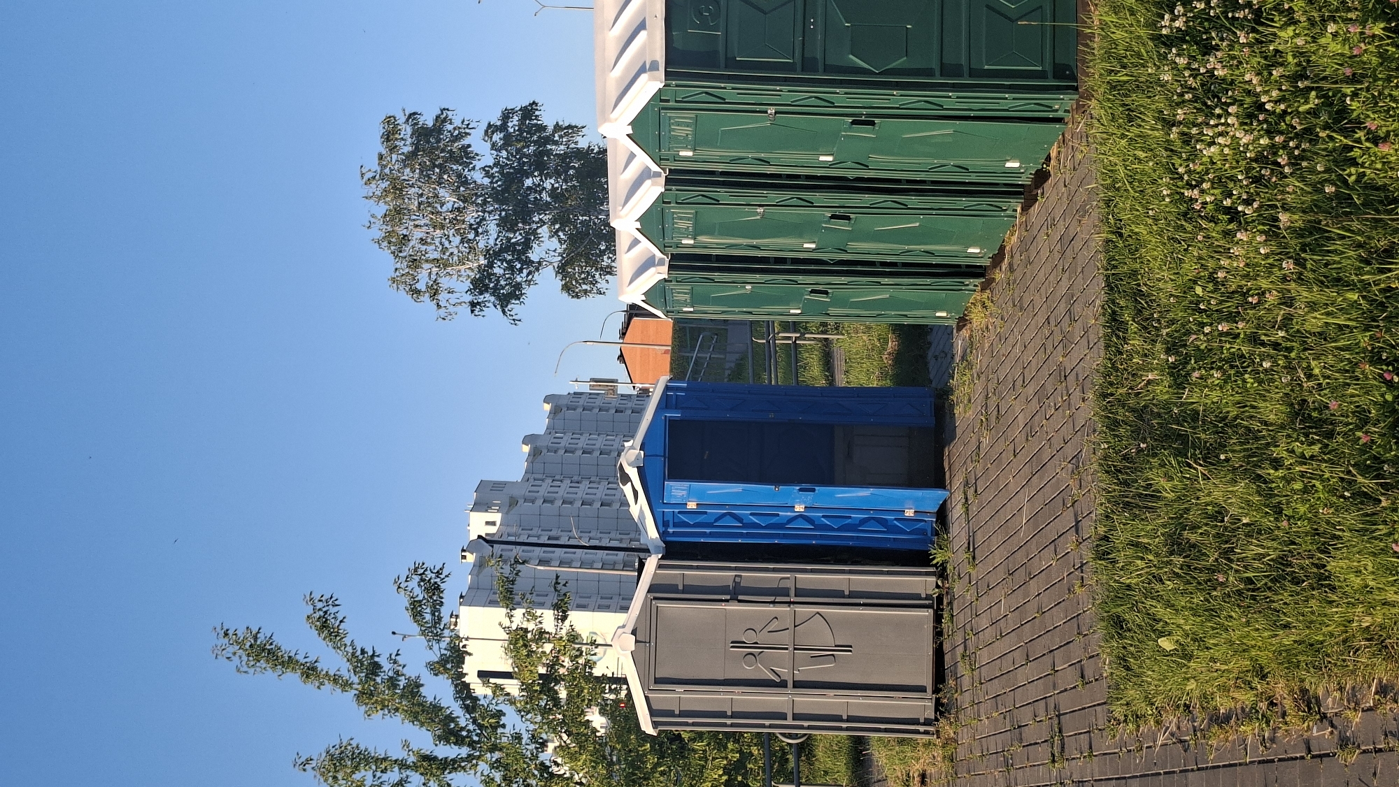 In the city of Smaliavičy, Belarus, an outdoor scene unfolds under a clear blue sky, bathed in the warm light of the sun. The foreground is dominated by a strip of vibrant green grass, dotted with small white clover flowers, suggesting a well-maintained public area. Behind the grass, a paved walkway made of grey interlocking bricks leads the eye towards a row of portable toilet units.
A single grey portable toilet stands to the left, adorned with a gender-neutral restroom symbol. Next to it, an open blue portable toilet invites entry, its door ajar. To the right of the blue unit, a line of dark green portable toilets stretches into the distance, their doors closed and uniform. The toilets are arranged in a neat row, suggesting a temporary setup for an event or a public facility.
In the background, several buildings rise against the horizon. A tall, multi-story white residential building with numerous windows stands prominently, its modern architecture contrasting with the utilitarian portable toilets. To its right, a smaller, brick-colored building is partially visible, hinting at a mix of urban development. Trees with lush green foliage are scattered throughout the scene, one tall, slender tree with sparse leaves situated between the buildings and the portable toilets, and another leafy tree framing the left side of the image. A streetlamp stands near the buildings, indicating a developed urban environment. The overall impression is one of a functional outdoor space within a Belarusian city.