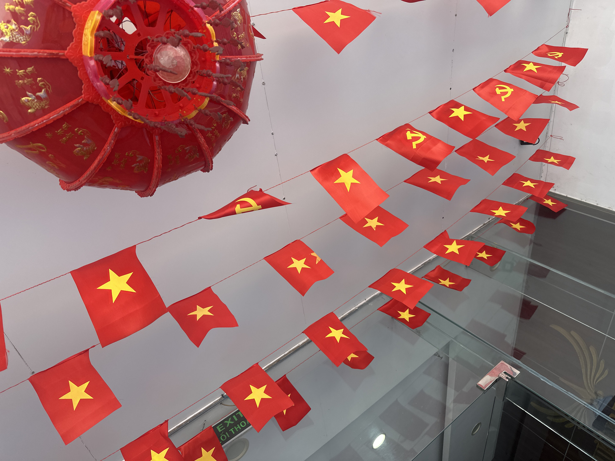 The image shows a decorated interior, possibly in Bac Nha Trang Ward, Vietnam, with red and yellow Vietnamese flags strung across the ceiling. A large red lantern hangs prominently on the left. The interior features white walls and a glass-enclosed area is visible.
