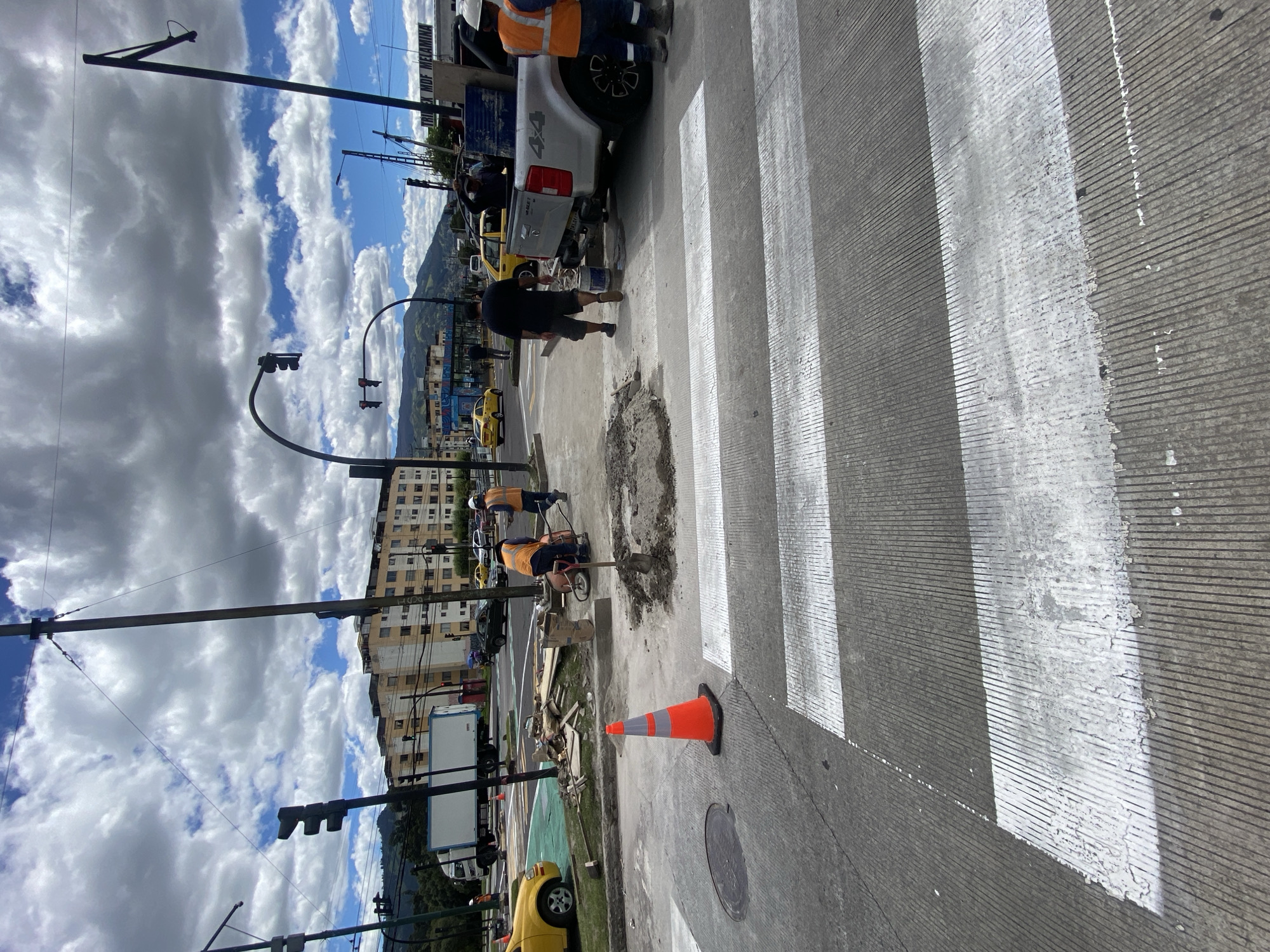 The image shows a street scene in Quito, Ecuador, focusing on a crosswalk under repair. A team of workers is present, with one applying a liquid substance (possibly paint or sealant) to the road surface near a damaged area. Another worker operates a small machine, possibly a grinder or mixer, nearby. Several vehicles are parked nearby, including a pickup truck. An orange traffic cone is positioned on the side of the road to alert passersby. The crosswalk is marked with white stripes that show signs of wear and tear. In the background, there are apartment buildings, power lines and traffic lights. The sky is partly cloudy. In the distance, a mountain range is visible.