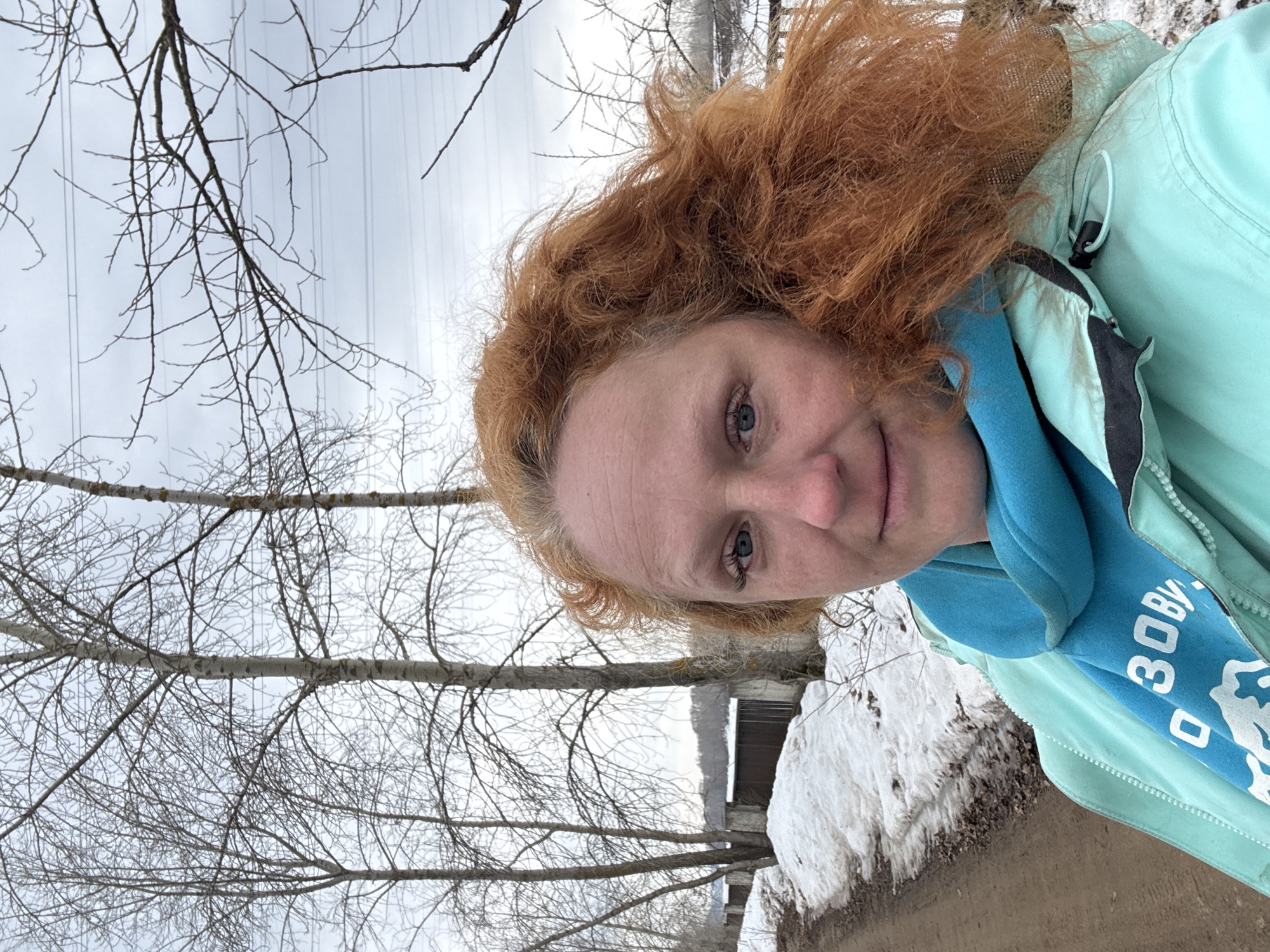 A fair-skinned woman with red curly hair is pictured in an outdoor setting. She wears a light green jacket and a blue scarf with white lettering in Russian. The background includes bare trees, a cloudy sky, patches of snow, and the corner of a building. The woman is situated in Chaikovsky, Russia.