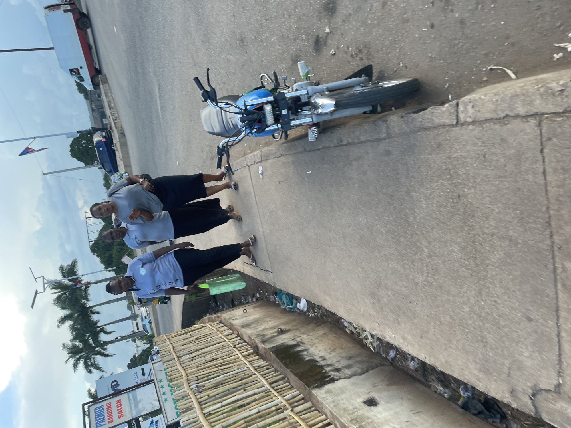 Three women are walking on a sidewalk next to a busy road. The woman in the foreground is wearing a light blue t-shirt and black pants, carrying a green shopping bag. The woman next to her is wearing a gray t-shirt and black skirt, with her left hand raised in a gesture. The third woman is slightly behind them, also wearing a gray t-shirt and black pants. They are walking away from the camera. A parked motorcycle is visible on the right side of the sidewalk. In the background, palm trees, a white van, and a blue car are visible. The sidewalk is cracked and shows signs of wear. A drainage ditch filled with debris runs alongside the sidewalk on the left. The sky is cloudy.