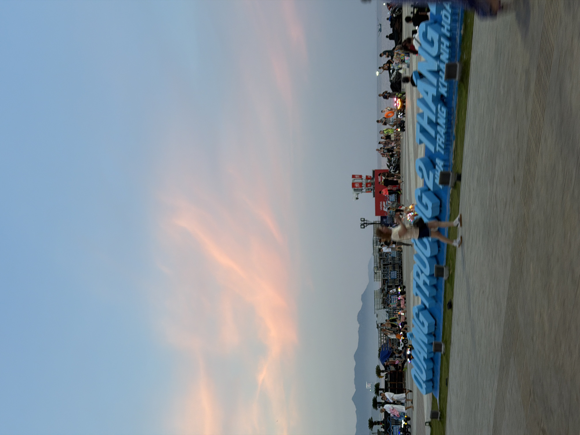 This is a full-shot, eye-level, vertical, outdoor photograph taken at sunset in Nha Trang, Vietnam. In the foreground, a woman in a tan shirt and dark shorts walks from right to left. Behind her, a large, light blue, three-dimensional sign reads "QUANG TRƯỜNG 2 THÁNG NHÀ TRANG KHÁNH HÒA." The sign is set on a patch of grass, with a paved walkway in front of it. In the background, a crowd of people stand near various tents and structures. Some individuals are interacting with small vehicles. In the distance, mountains are visible against the dusk sky, which is a gradient of pale blue to soft pink. The sea is visible behind the crowd, with a small boat on the water.