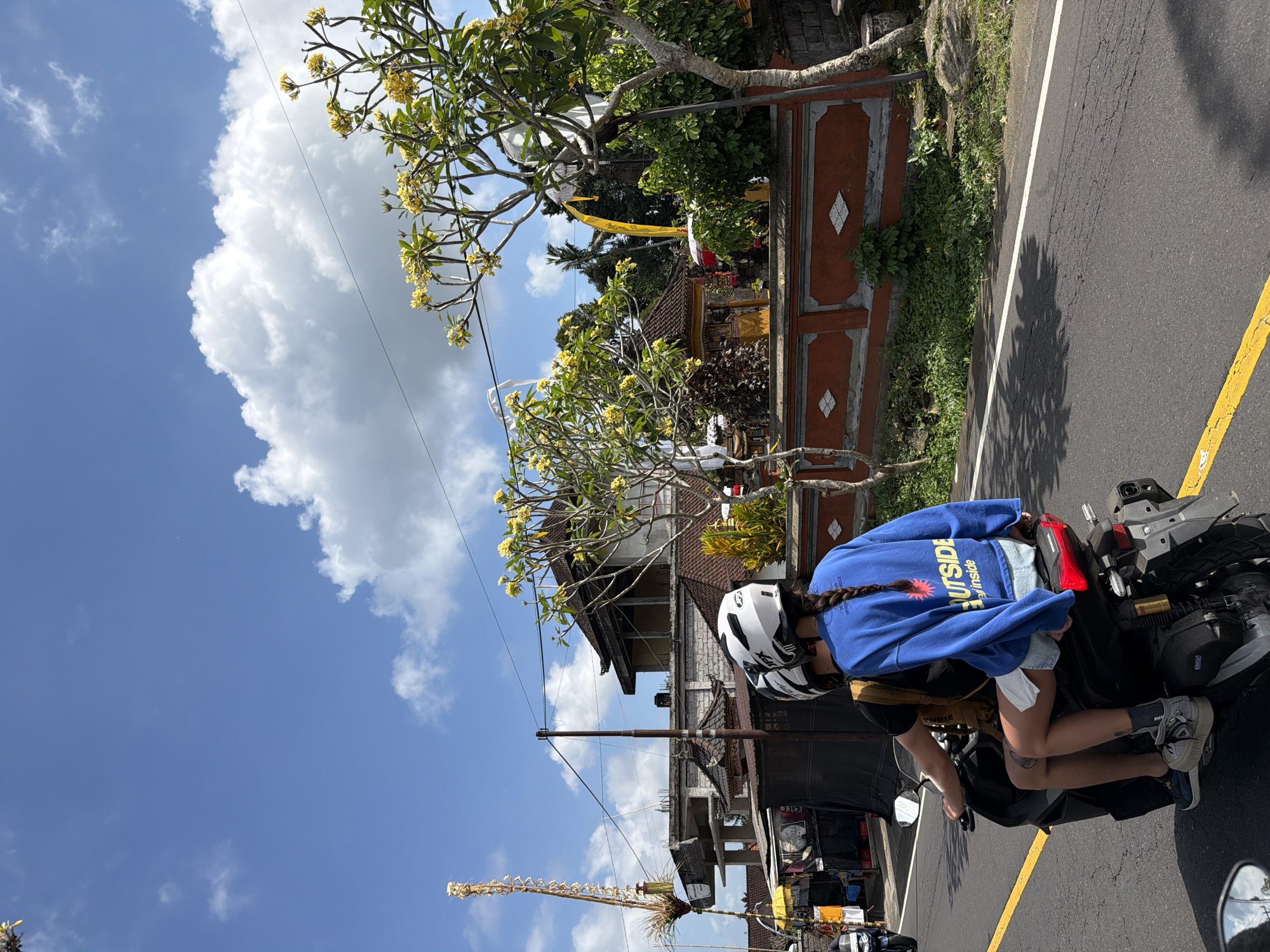 In Tampaksiring, Indonesia, two individuals wearing helmets ride a motorcycle on a paved road under a bright blue sky with scattered white clouds. The person in the back, wearing a blue sweatshirt and denim shorts, faces away from the camera, her braid visible. The person in front, wearing a black t-shirt and a backpack, also faces away. Behind them, a low, reddish-orange wall with diamond-shaped accents borders a lush green area and several buildings with traditional tiled roofs. Yellow frangipani trees with white and yellow flowers are visible, along with a tall, decorative bamboo structure on the left. The road is marked with yellow and white lines.