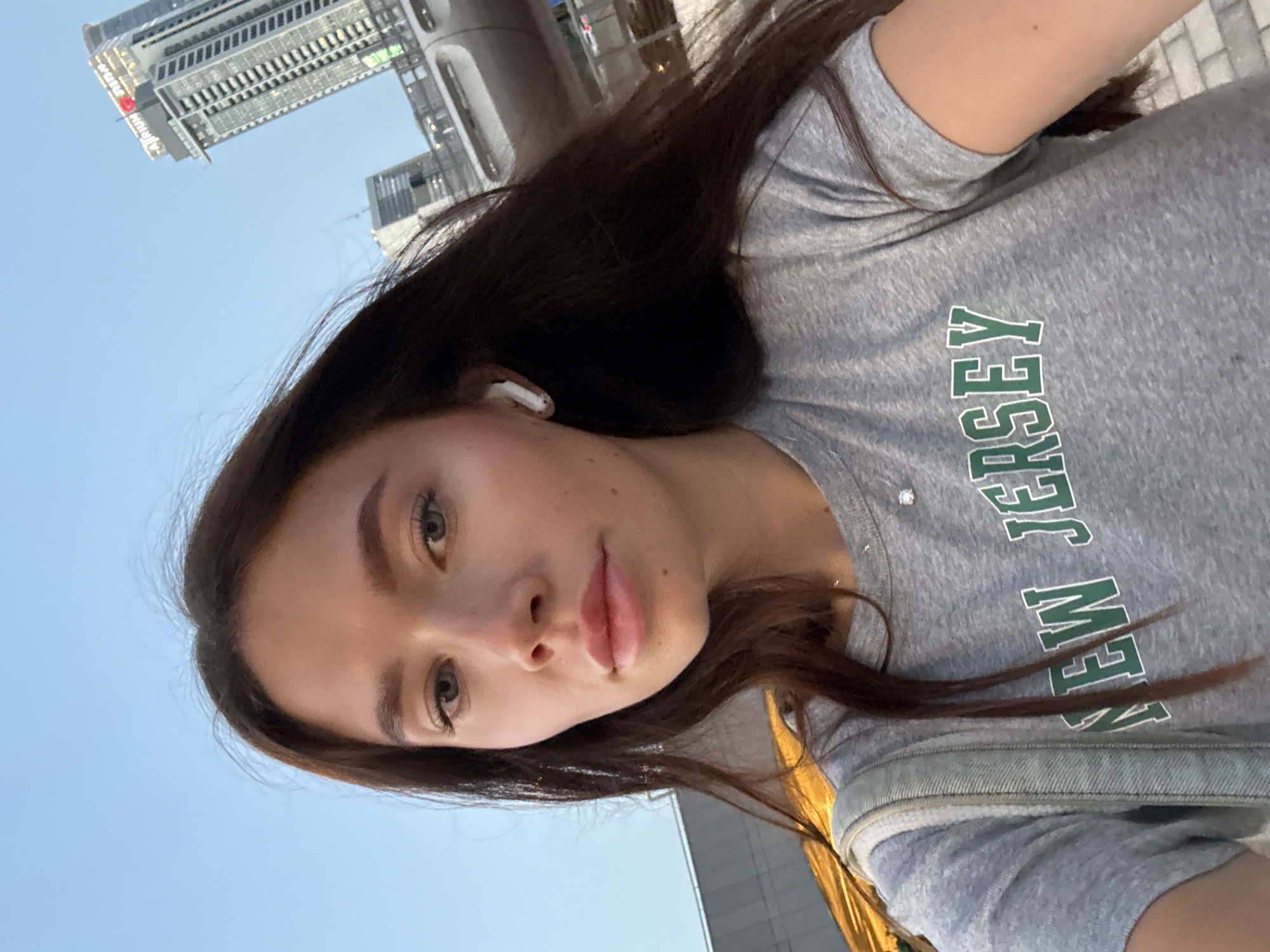 A young woman is depicted in a self-portrait, taken from a slightly upward and tilted perspective. She has long, dark brown hair and light-colored eyes, with a calm, neutral expression. A white earbud is visible in her right ear. She is wearing a grey t-shirt with the words "NEW JERSEY" printed across the front in green letters with a white outline. A light-colored denim strap, possibly from a bag or jacket, is visible over her shoulder.
The setting is an outdoor urban environment under a clear, light blue sky, suggesting daytime, possibly late afternoon. In the background, several modern, tall buildings define a city skyline. On one of the prominent buildings in the distance, the red text "AZRIELI" is discernible, which strongly indicates the location is Tel-Aviv, Israel, specifically near the Azrieli Center. The overall scene conveys a casual moment captured in a bustling city.