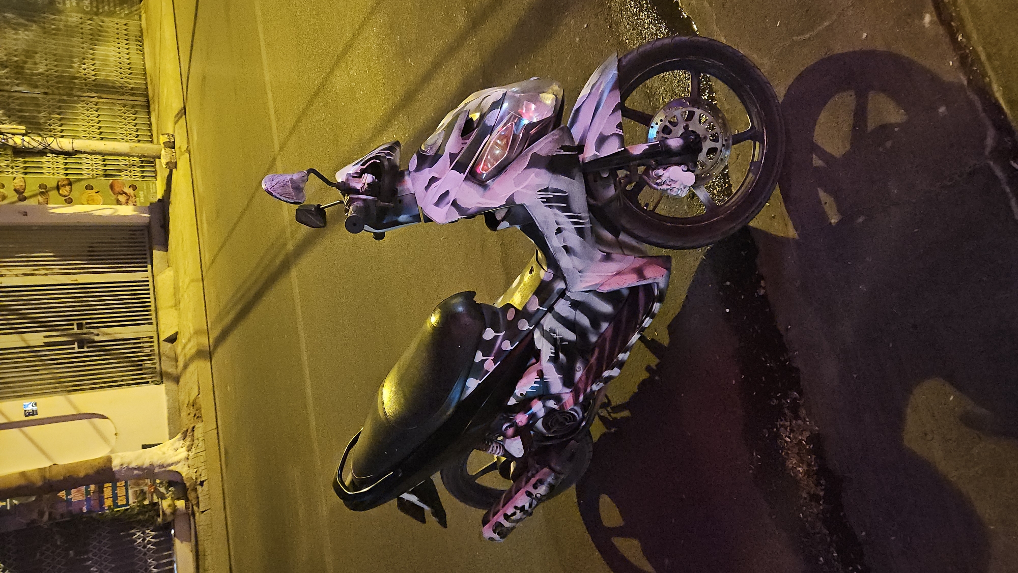 A motorcycle with custom pink and black paint stands parked on a street. The location is likely within the city of Bắc Nha Trang, Vietnam. The road surface is paved and dimly lit, possibly by streetlights. The motorcycle's front wheel is visible, and its shadow is cast onto the road. In the background, there is a building with a closed metal gate or shutter.