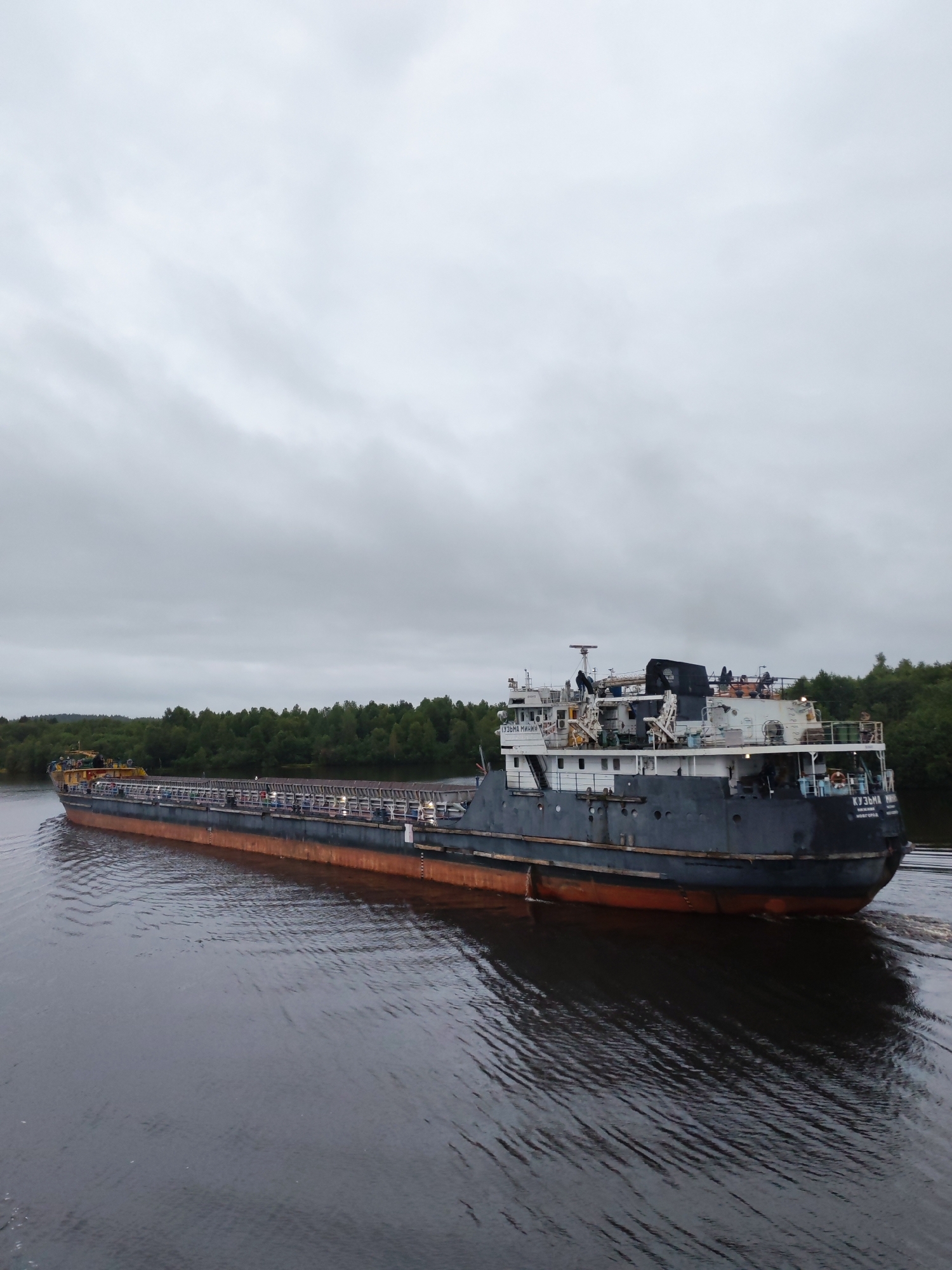 A barge is navigating a wide river under a cloudy sky. The barge is long and appears to be carrying cargo, indicated by the numerous cylindrical containers stacked on its deck. The vessel has a dark hull with an orange waterline. The superstructure of the barge is white and gray, featuring various equipment and structures, including cranes and cabins. Several figures are visible on the deck of the barge. The riverbanks are lined with dense green trees. The water is calm with visible ripples, and the barge casts a dark reflection. The image is taken from a slightly elevated perspective, looking towards the barge and the distant treeline. The text "КУЗЬМА МИНИН" is visible on the side of the barge. The location is identified as Devyatiny, Russia.