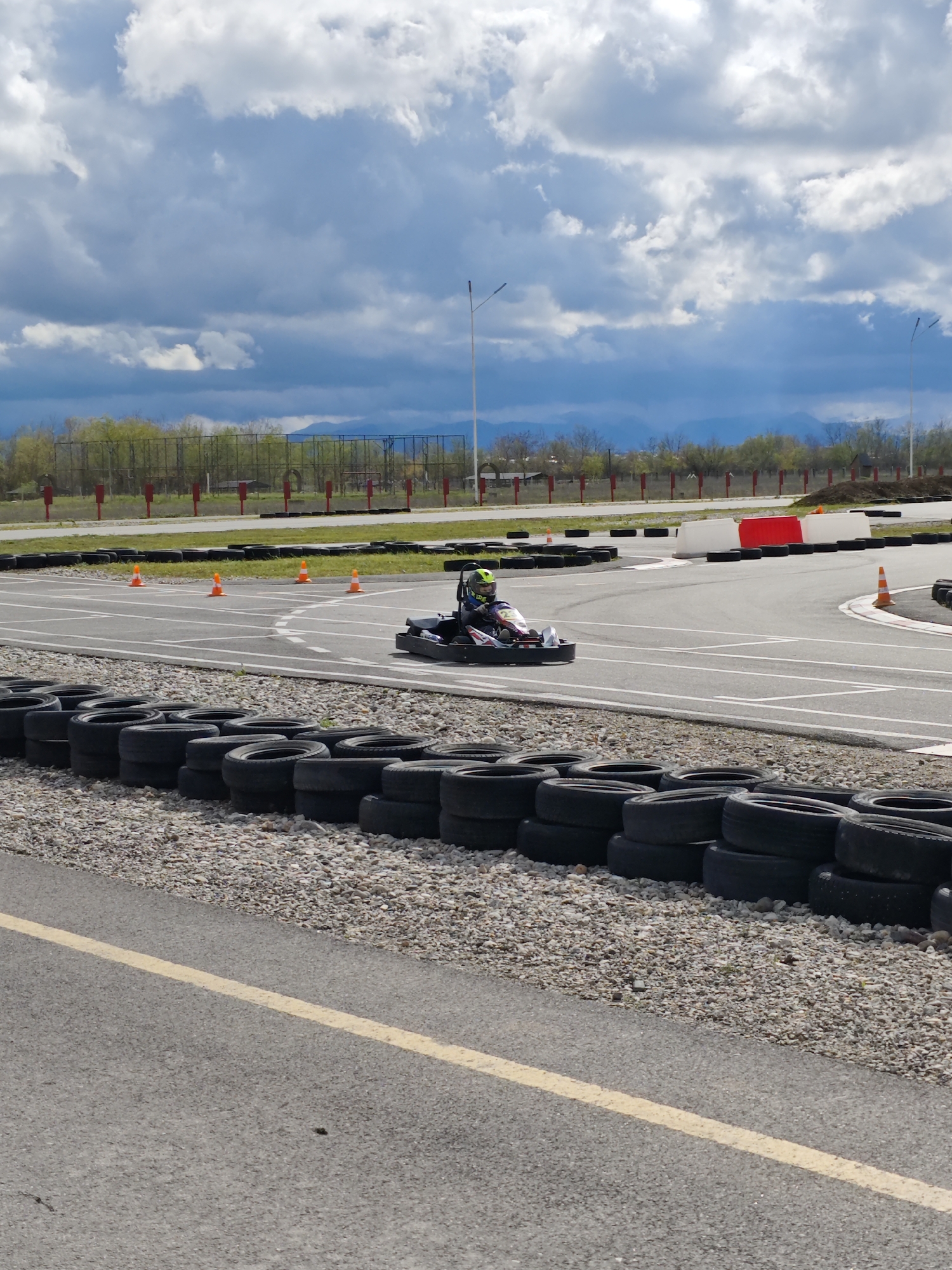 The image depicts a go-kart track, likely in Kishpek, Russia, based on geographical data. A go-kart with a driver wearing a helmet is visible on the track, navigating a turn marked by orange cones. The track is bordered by stacks of tires and a gravel area. In the background, a chain-link fence, trees, and mountains are visible under a cloudy sky. Two utility poles stand in the background. The scene appears to be an outdoor racing facility.