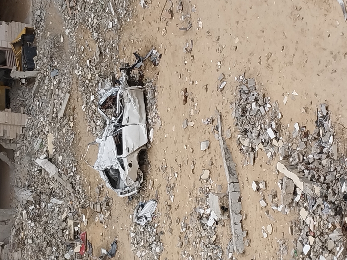 The image depicts a scene of extensive destruction in Khan Yunis, Palestinian Territory. Centrally positioned is a severely damaged white four-door sedan, its roof caved in, body panels extensively crumpled, and the front end mangled. The vehicle rests on light brown sandy earth, which is densely strewn with a wide array of construction debris, including concrete fragments, rebar, and shattered masonry. In the background, remnants of at least one heavily damaged building are visible, characterized by crumbling concrete block walls and large piles of rubble. A detached piece of white vehicle bodywork lies on the ground to the immediate left of the wrecked sedan. No individuals are present in the scene.