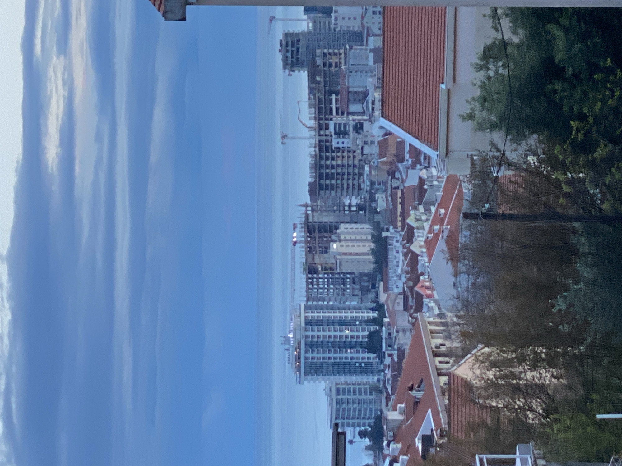 The image depicts an overview of a cityscape. On the left side of the image is a sky filled with striated clouds. Adjacent to the sky is the coastline of a body of water. The central area is populated with a dense urban landscape, featuring buildings of varying heights, some under construction, with cranes visible. The buildings are located near the city of Budva, Montenegro. Red-tiled rooftops, trees, and other vegetation are interspersed among the buildings. On the right side is green trees.