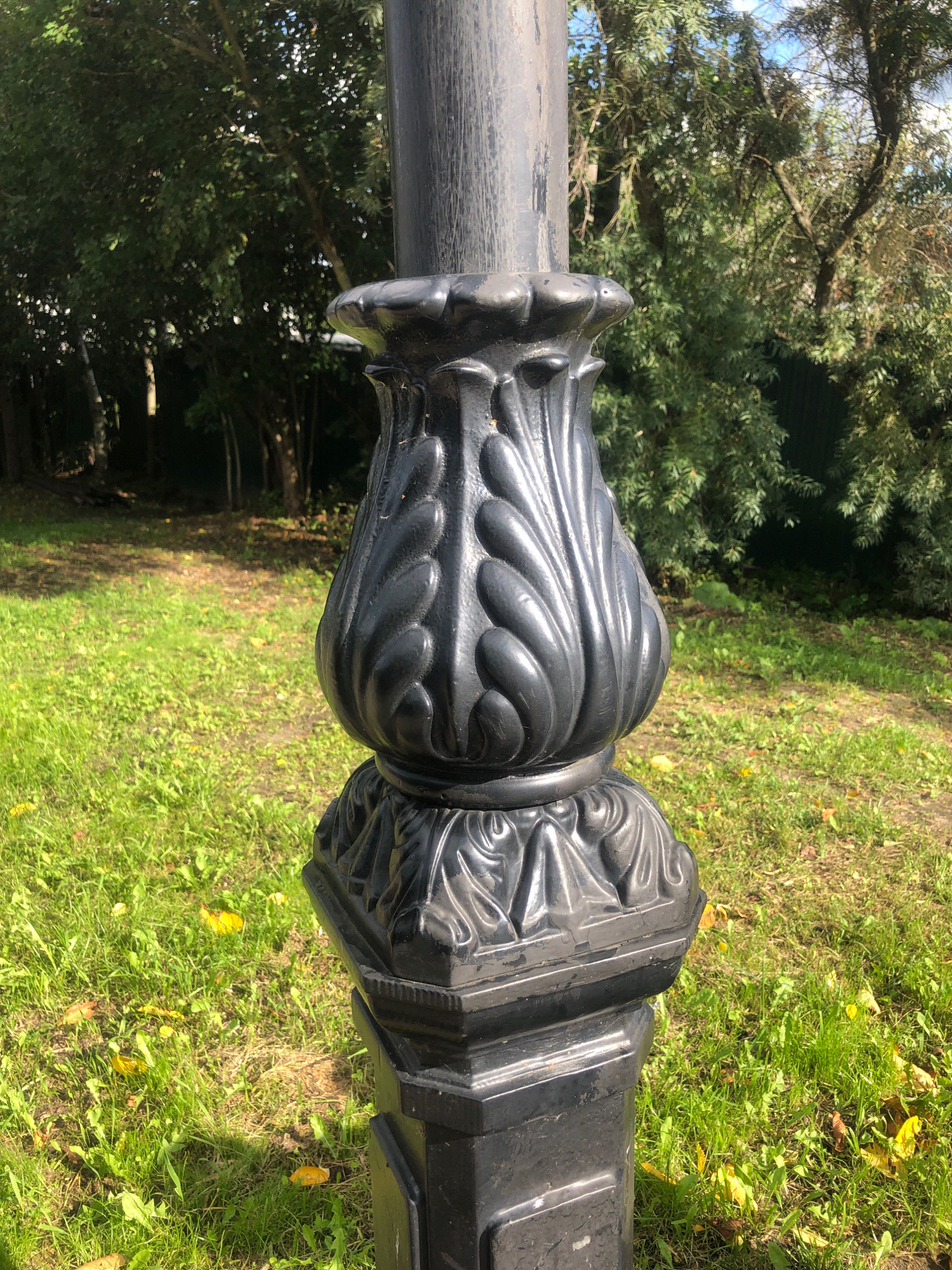 A black ornate lamppost stands in a grassy area with scattered yellow leaves. The lamppost has a decorative base with acanthus leaf motifs and a fluted upper section. Behind the lamppost, trees and foliage are visible, with a hint of blue sky. The scene appears to be in a park or public garden in Tutaev, Russia.